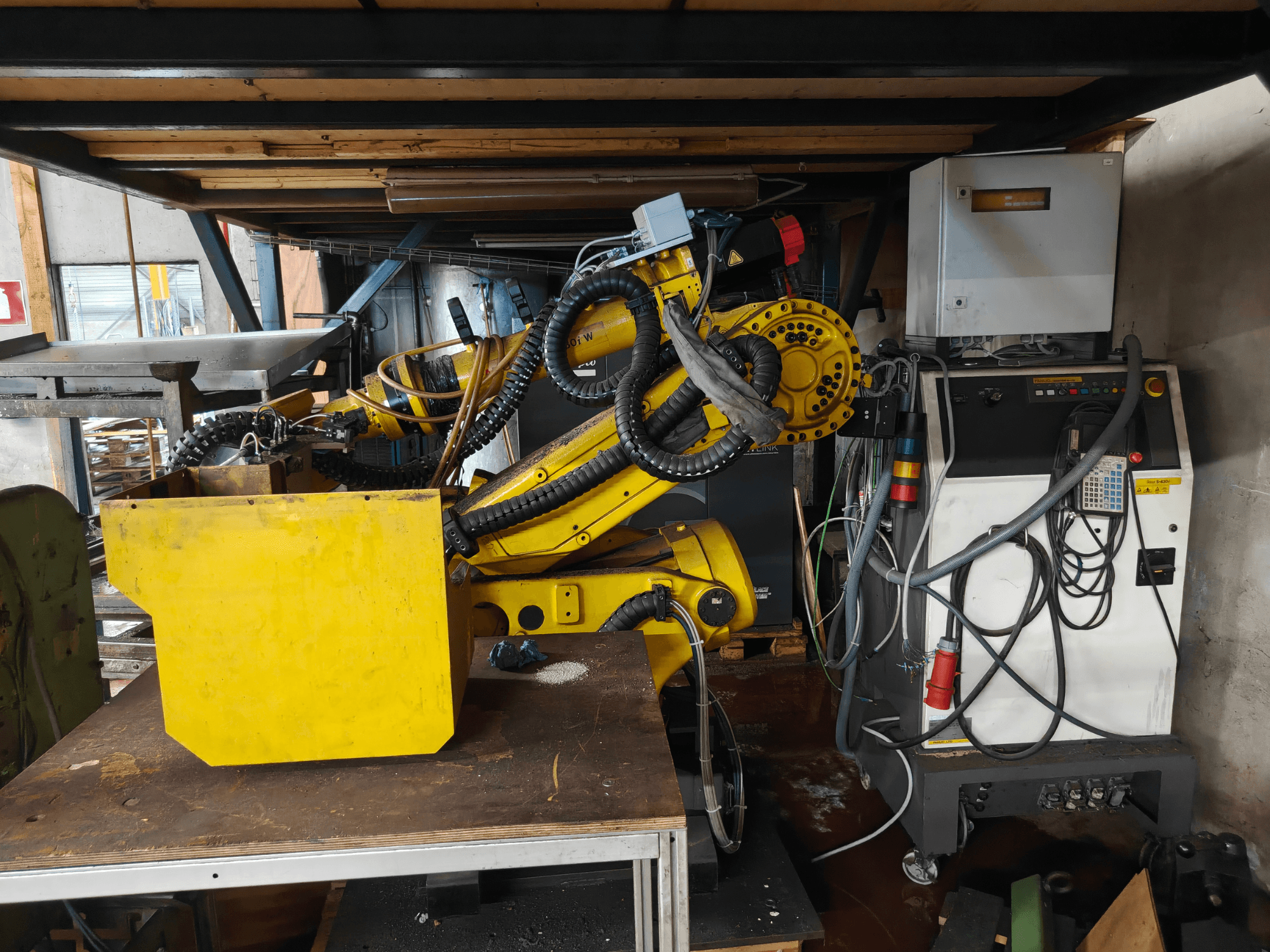 Vista frontale del braccio del robot FANUC S-430i W, di colore giallo, con pannello di controllo, cavi e un componente giallo su un banco da lavoro.