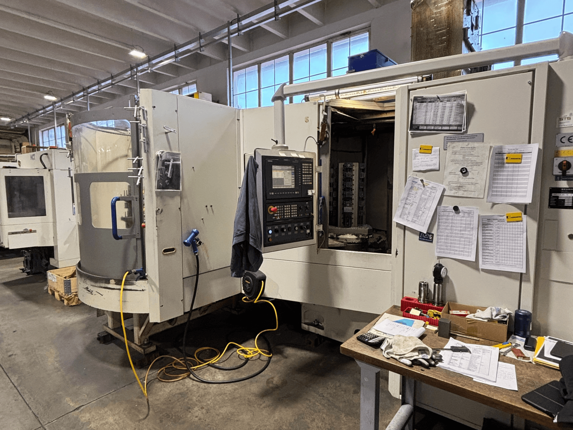 Macchina CNC in un'officina, vista frontale, con pannello di controllo e documentazione sul lato; utensili e materiali sul tavolo.