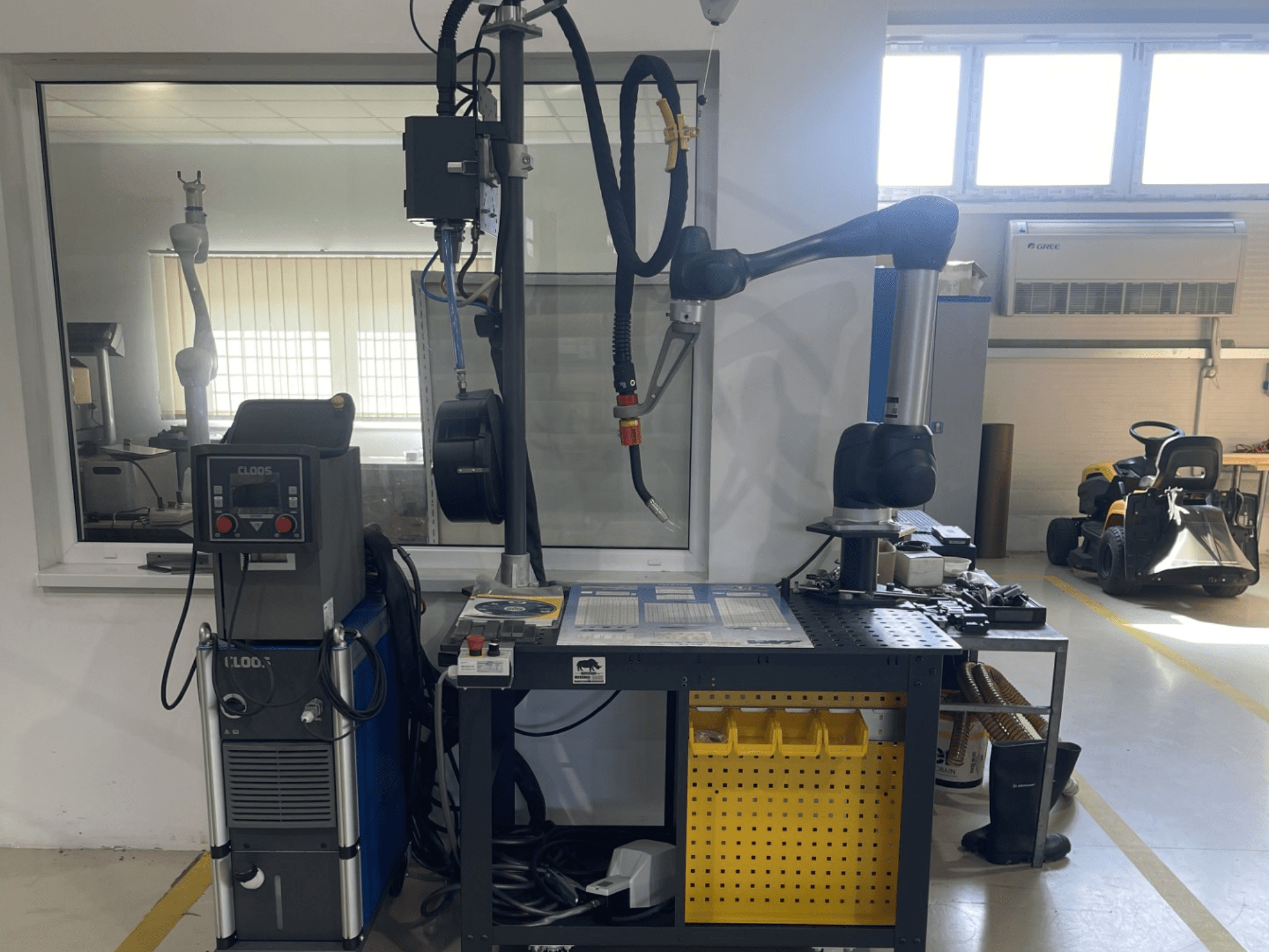 Robot collaborativo DOOSAN M1013 in un'officina, vista laterale con attrezzatura per la saldatura e utensili su un banco da lavoro.