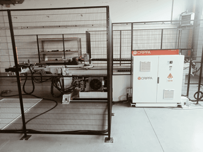 Macchina CRIPPA bianca dietro una gabbia di sicurezza, con tubi e componenti visibili, vista laterale, ambiente industriale.