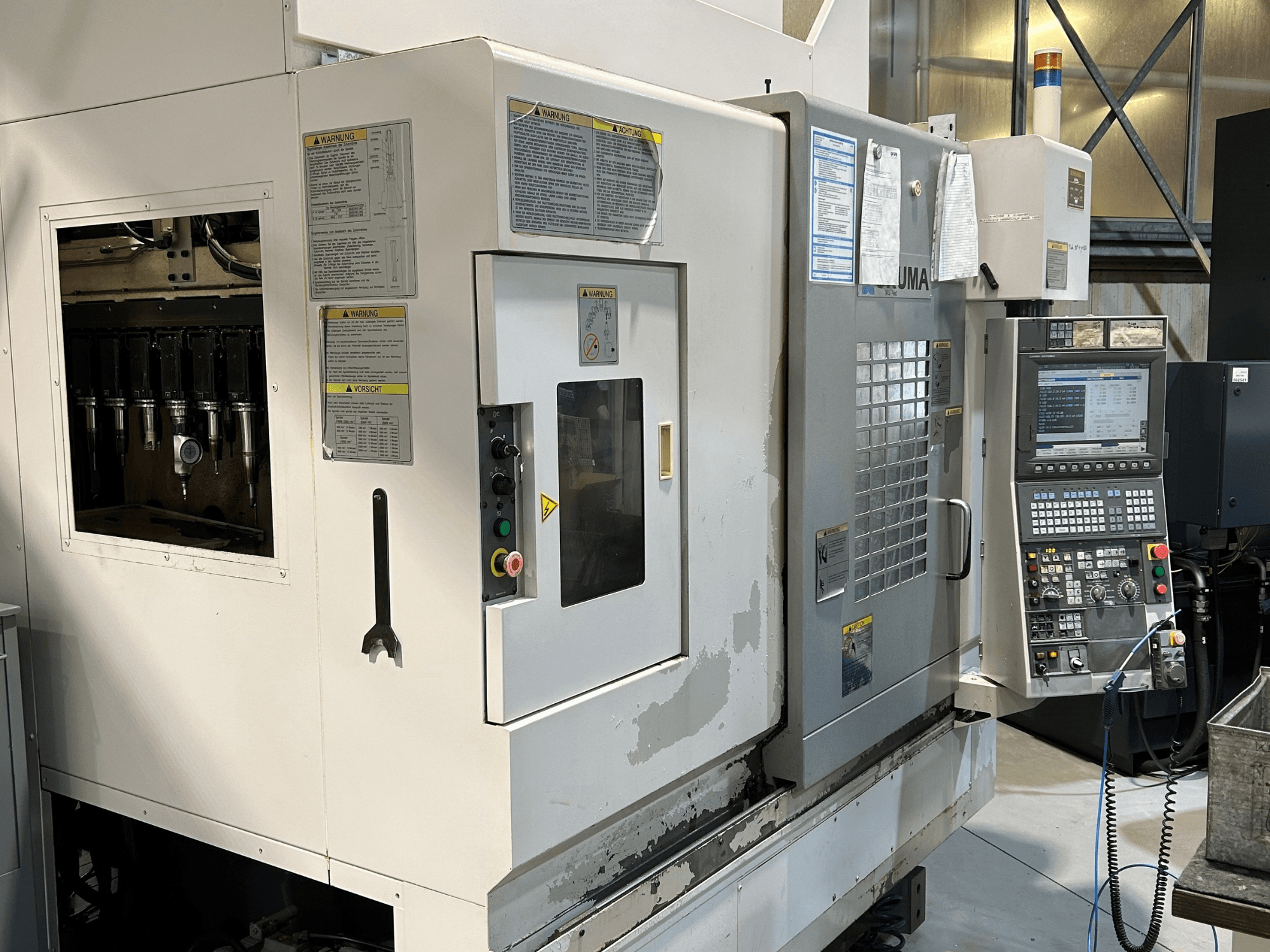 Vista frontale di una macchina CNC UMA con portautensili visibili, pannello di controllo, etichette di avvertenza e pulsanti operativi.