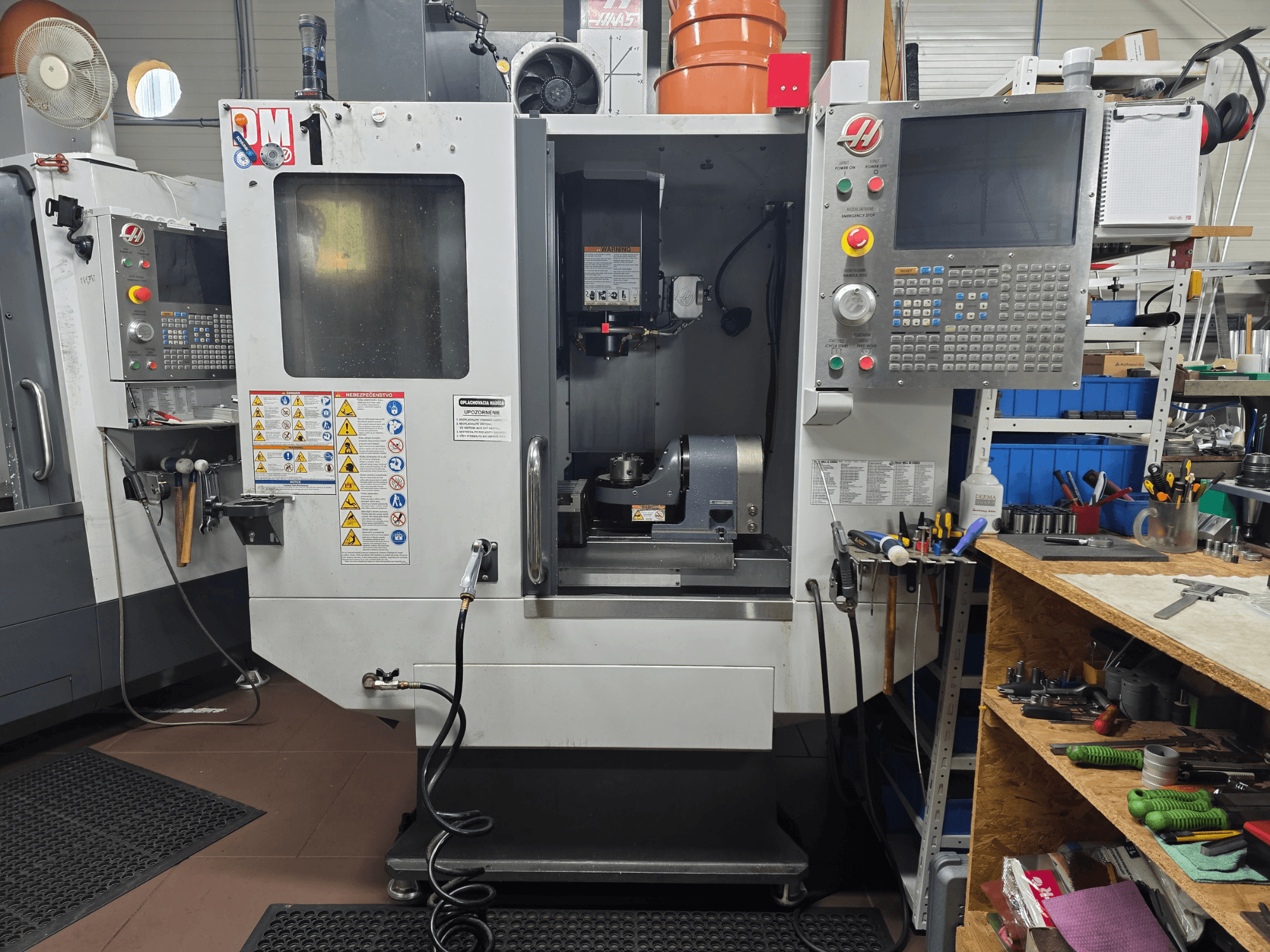 Macchina CNC Haas modello VF-2, vista frontale, che mostra i comandi, l'area del mandrino, le etichette di sicurezza e gli utensili su un banco di lavoro vicino.