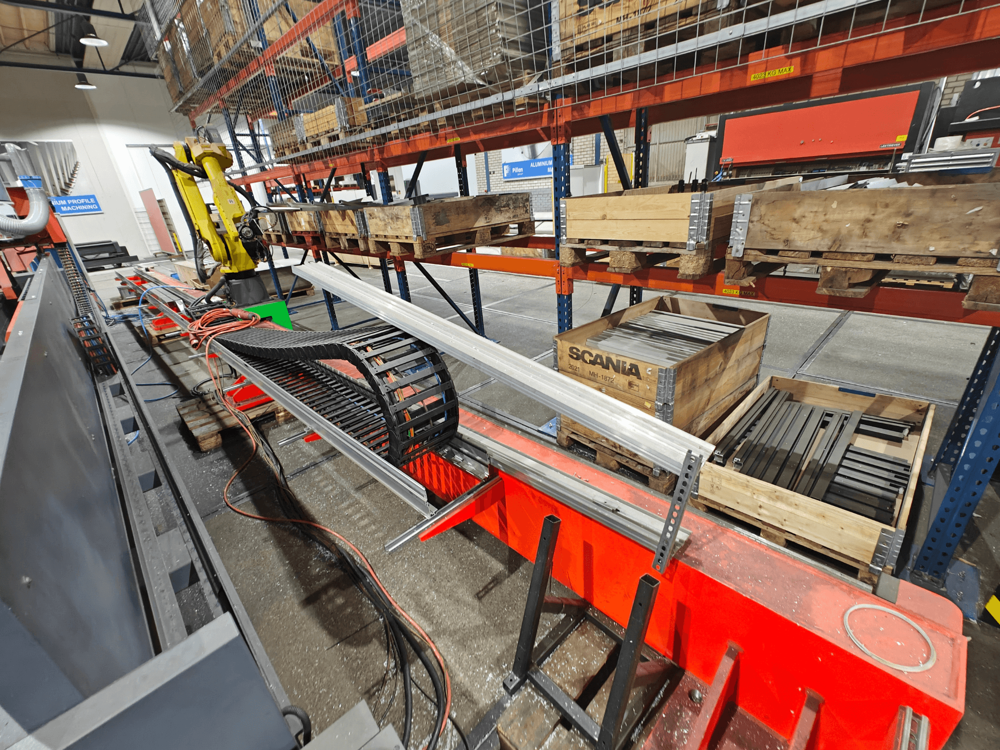 Braccio robotico su postazione di lavoro, cavi arancioni e neri visibili, con casse Scania e profili di alluminio in un ambiente industriale.