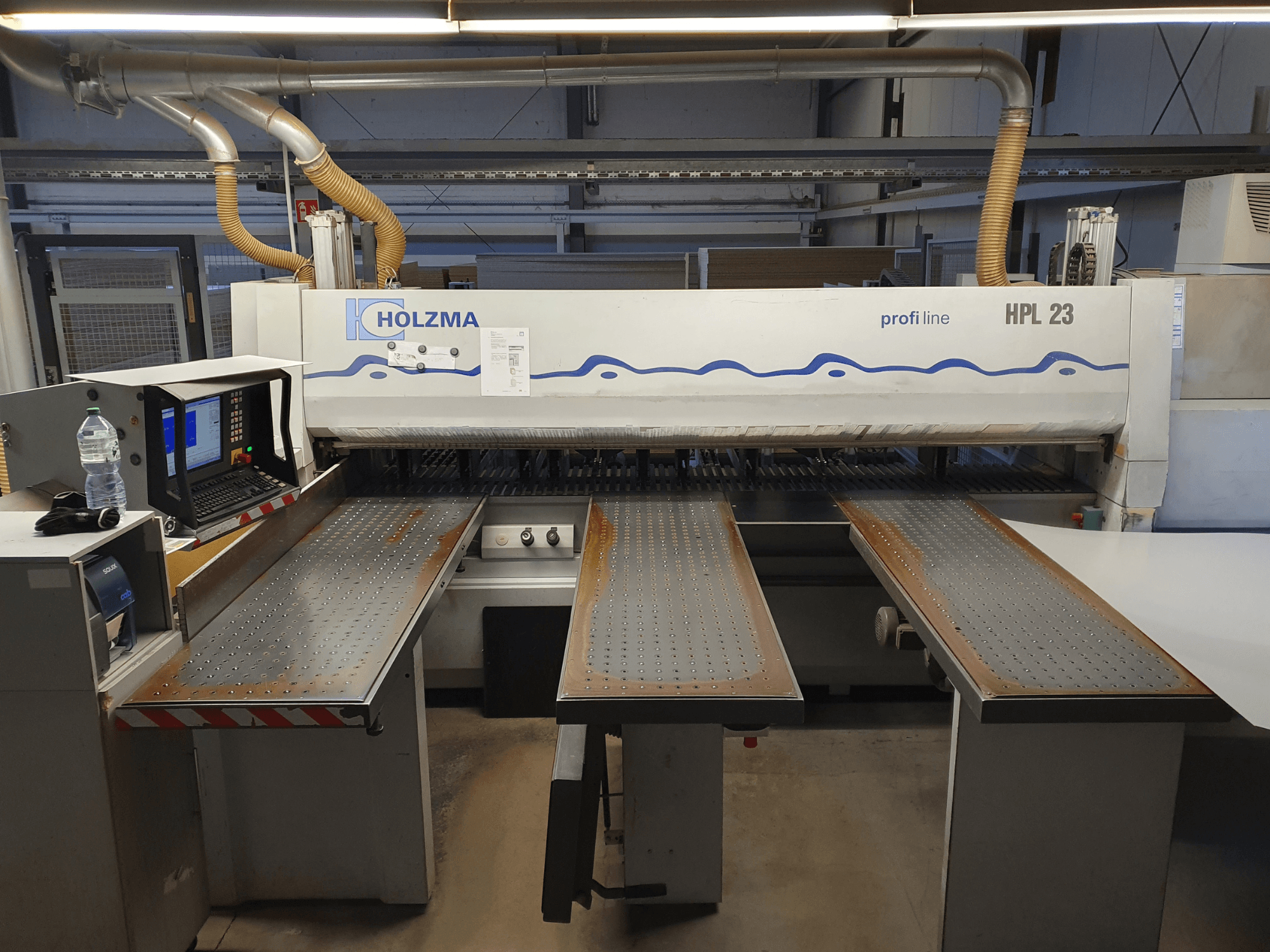 Macchina da taglio HOLZMA HPL 23 vista frontalmente, con tre tavoli perforati, terminale del computer e bottiglia d'acqua nelle vicinanze.