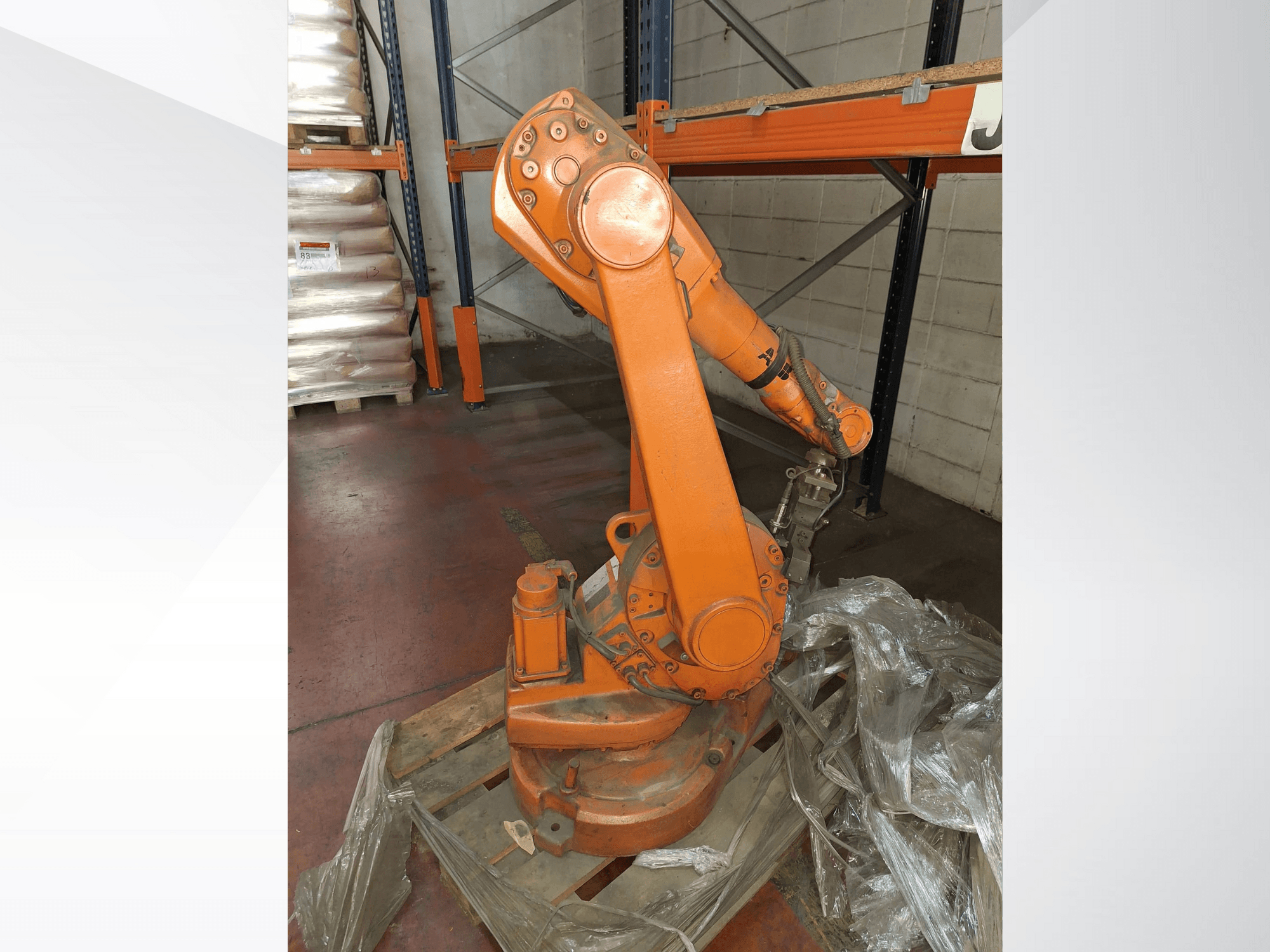 Braccio robotico industriale di colore arancione, vista frontale su pallet di legno, con cavo collegato e involucro protettivo in plastica intorno alla base.