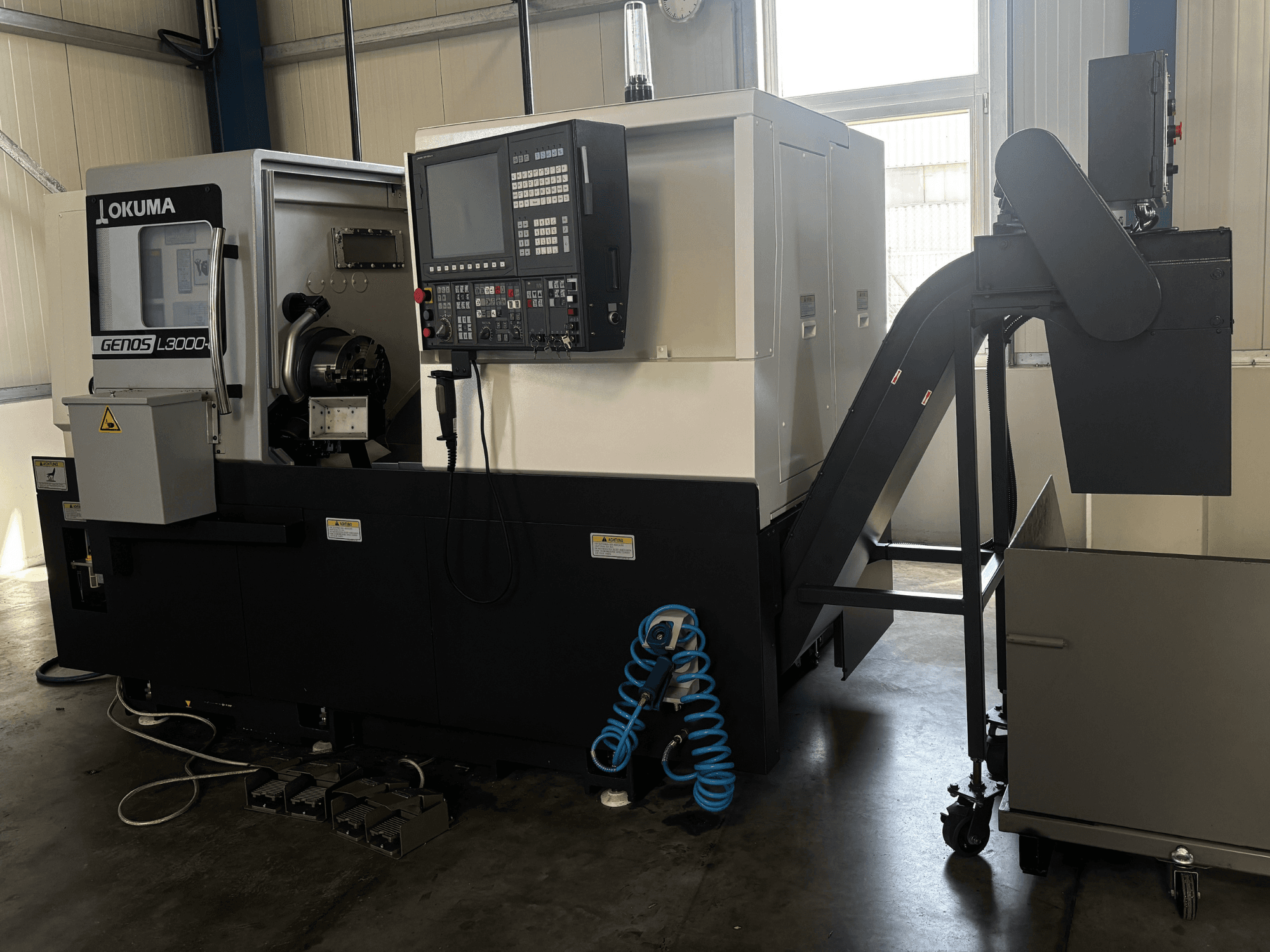 Tornio orizzontale Okuma GENOS L3000-e, vista frontale, con pannello di controllo, trasportatore e zona del mandrino visibile.
