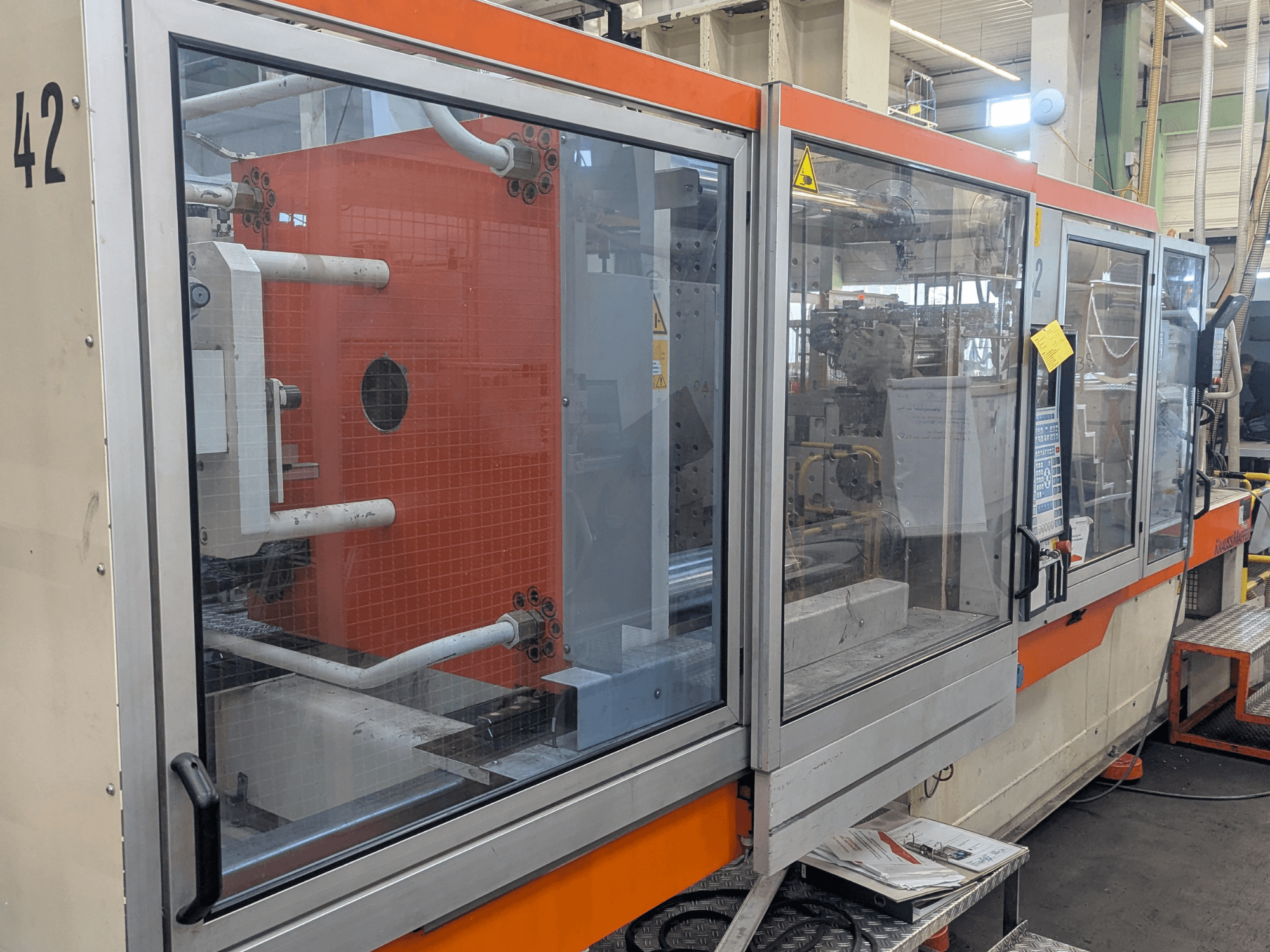 Pressa a iniezione idraulica Krauss Maffei KM 350-3000, vista laterale che mostra i comandi e i componenti interni dietro il pannello di vetro.