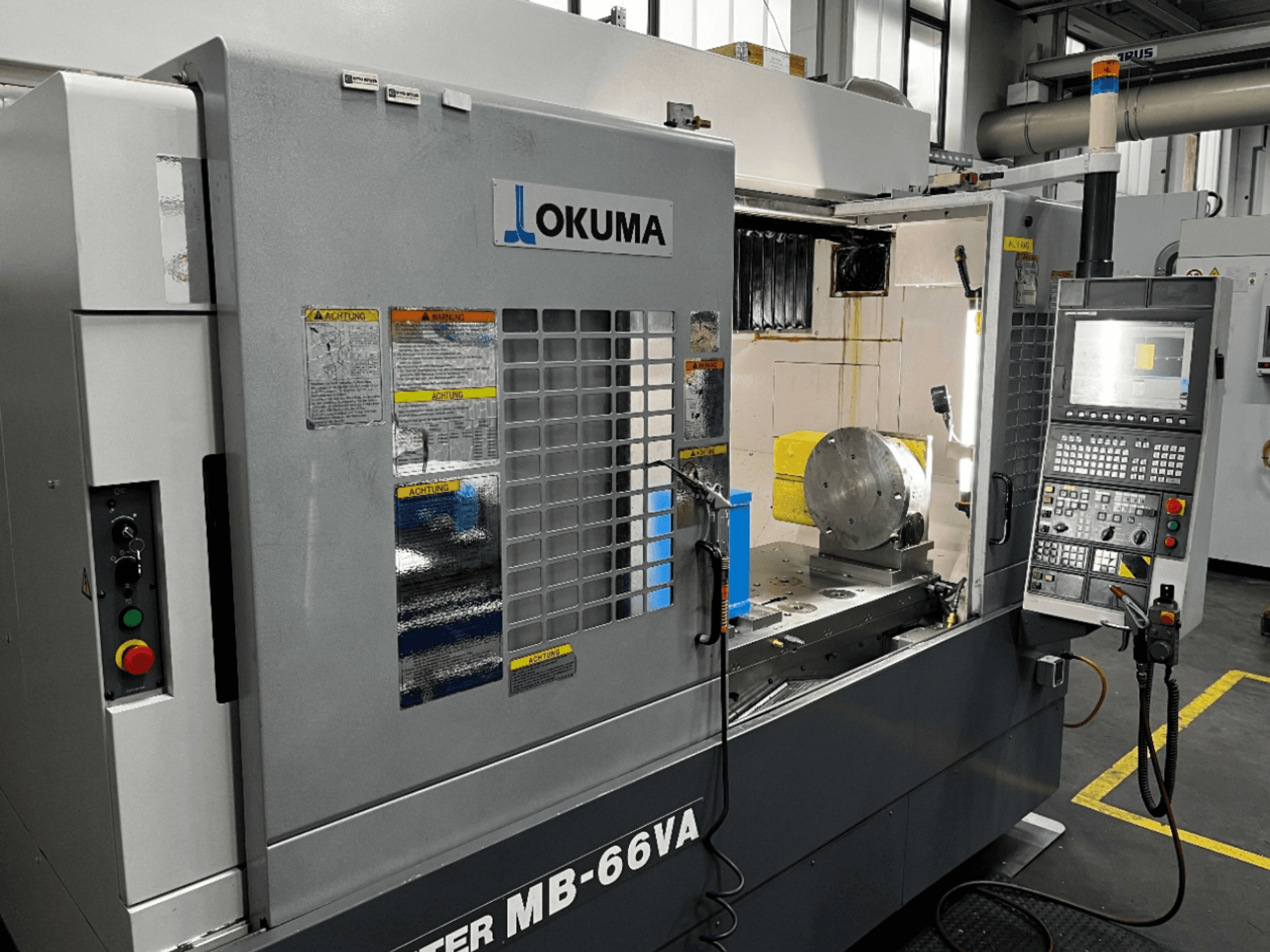 Centro di lavoro verticale Okuma MB66-VA vista frontale che mostra il pannello di controllo, il pezzo da lavorare e l'interno della macchina con le etichette di sicurezza.