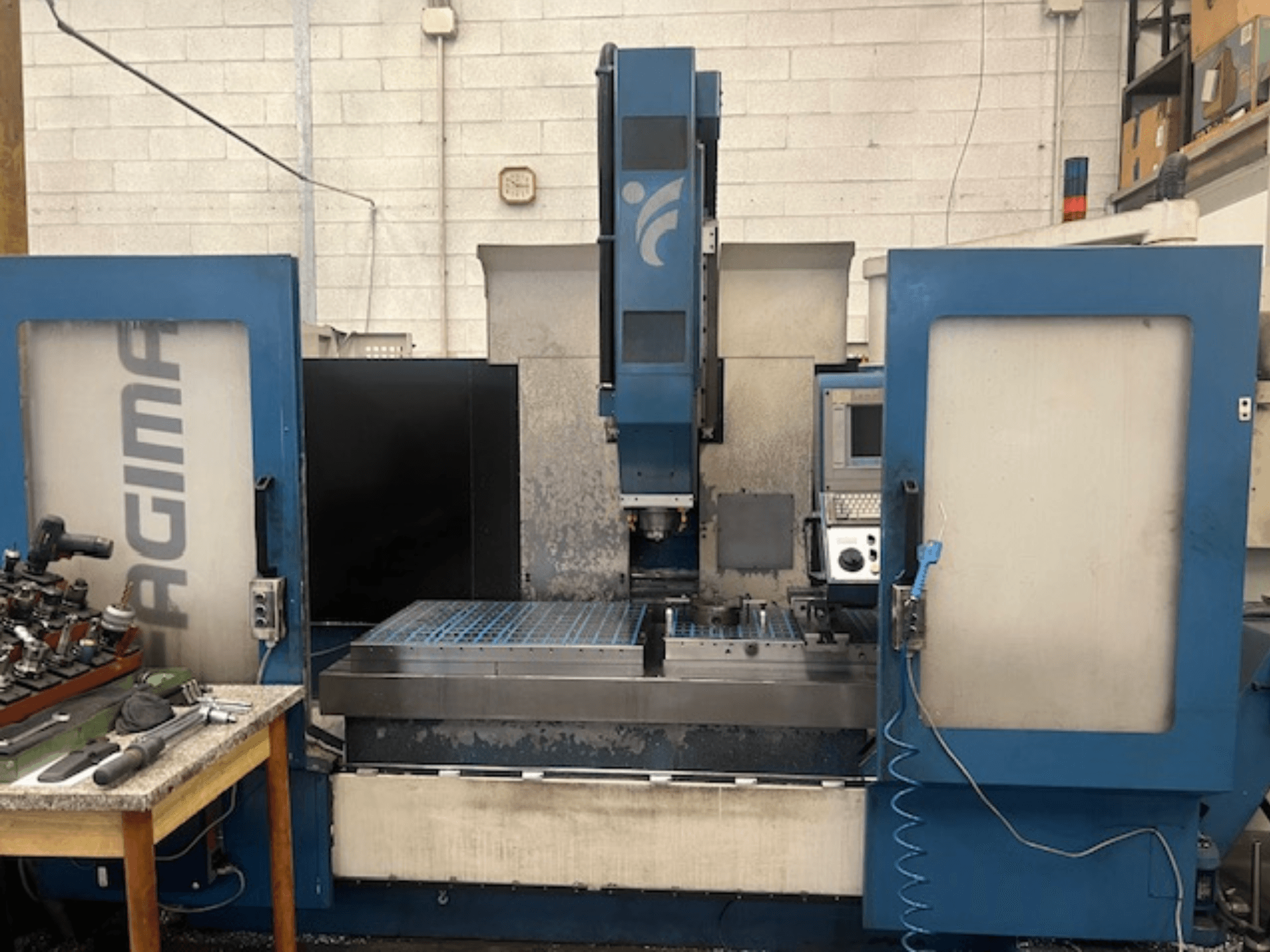 Vista frontale di una macchina CNC JAGIMA con un'ampia area di lavoro in metallo, un pannello di controllo visibile e utensili su un tavolo vicino.