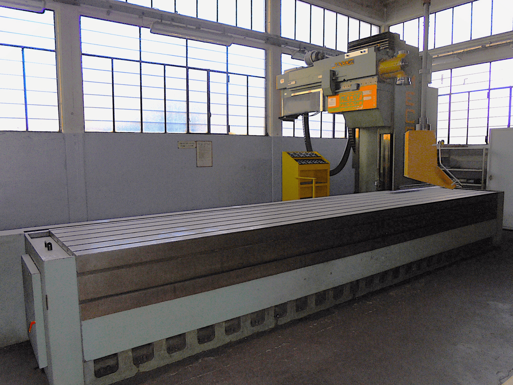 Fresatrice verticale di marca sconosciuta in una fabbrica, vista laterale, dotata di un grande tavolo con superficie scanalata e pannello di controllo.