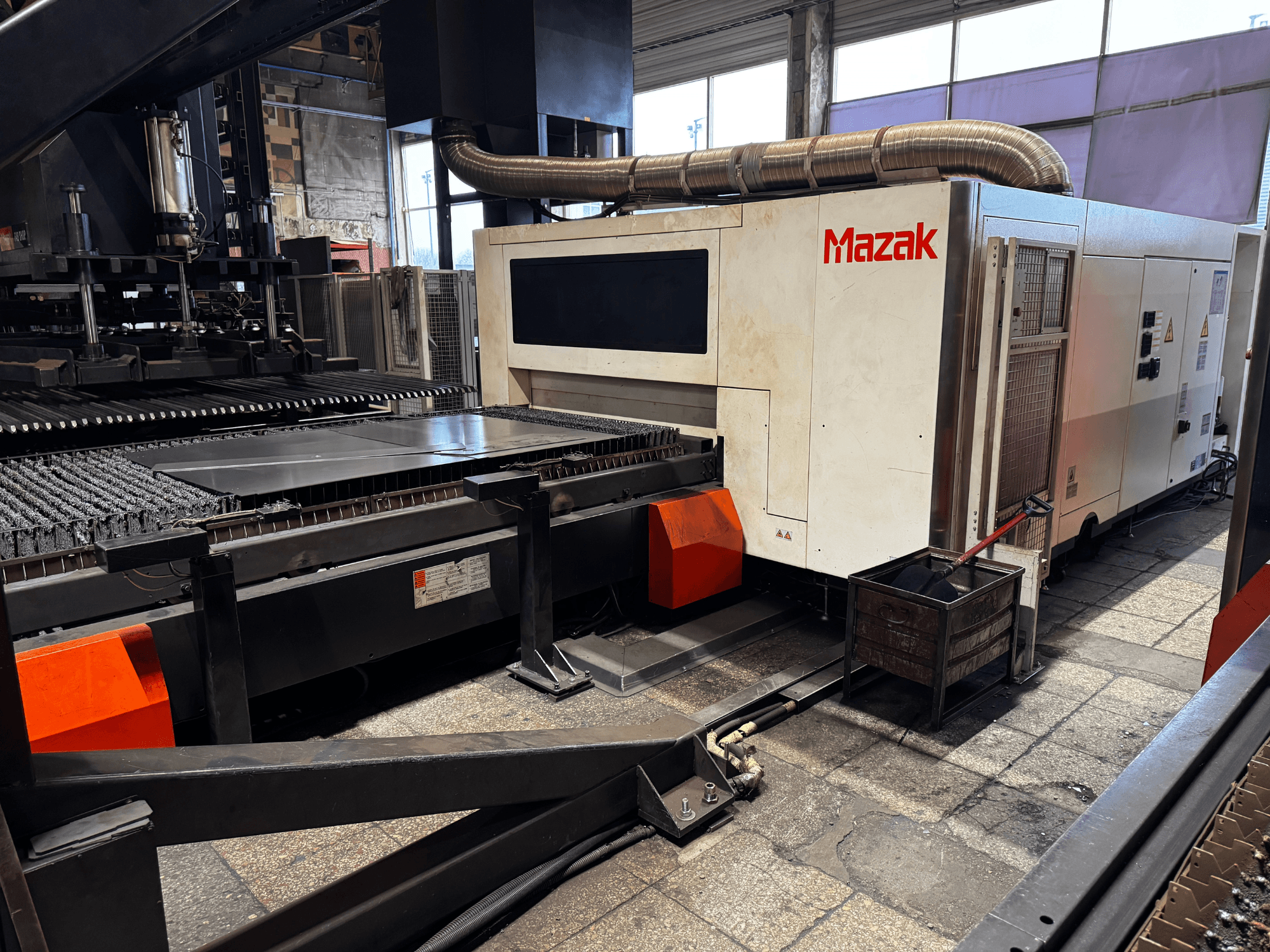 Macchina da taglio laser Mazak in un'officina, vista laterale che mostra il pannello di controllo, il trasportatore e un contenitore di raccolta.