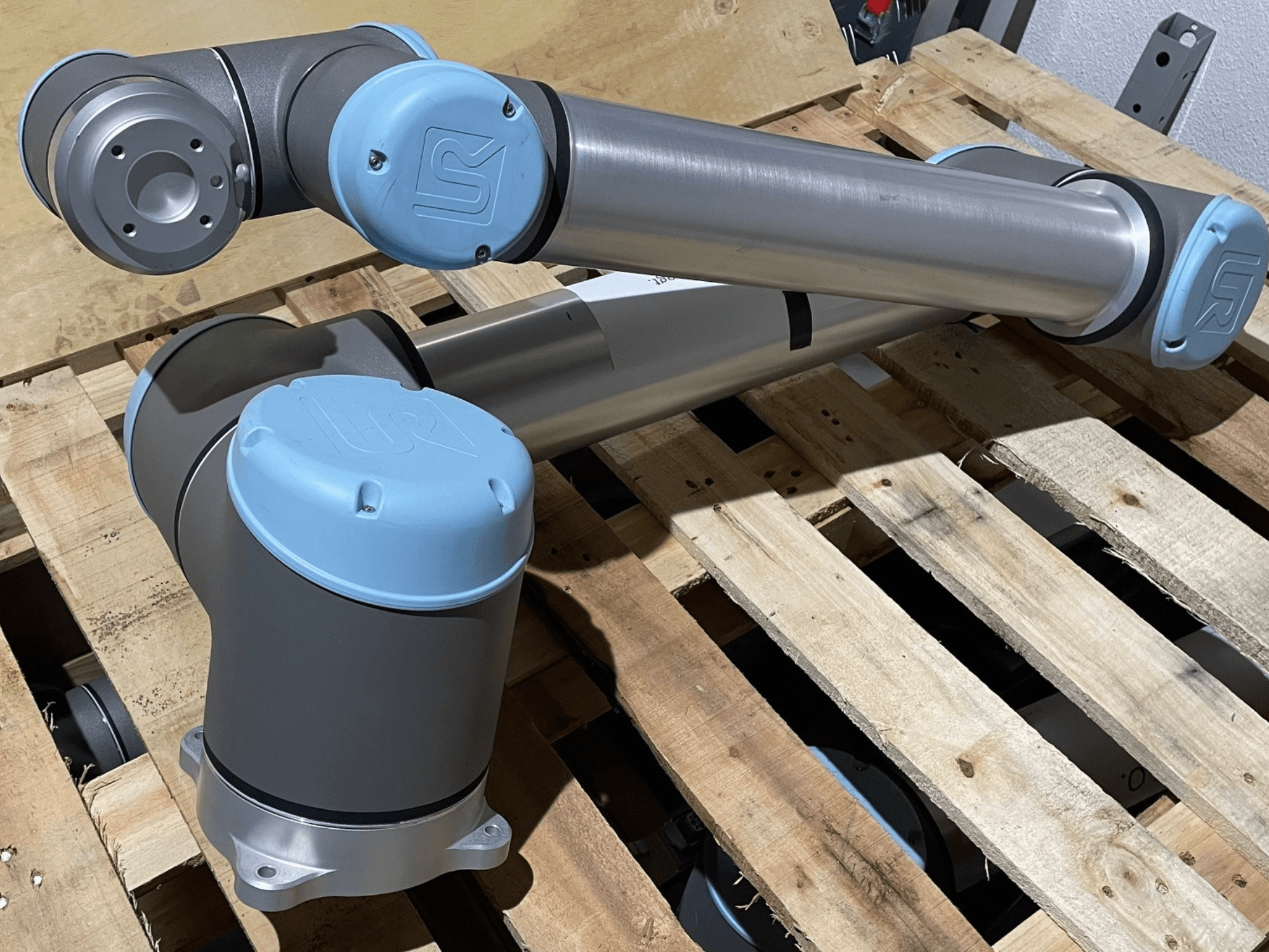 Braccio robotico argentato con terminali blu, vista frontale, con articolazioni e dettagli meccanici, appoggiato su un pallet di legno.