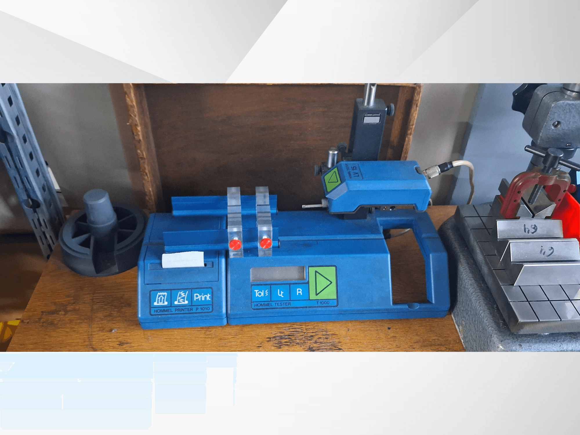 Stampante Hommel T 1000 blu e tester T 1 010 con pulsanti rossi, visti frontalmente su un banco di lavoro con blocchi metallici.