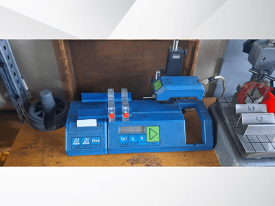 Stampante Hommel T 1000 blu e tester T 1 010 con pulsanti rossi, visti frontalmente su un banco di lavoro con blocchi metallici.