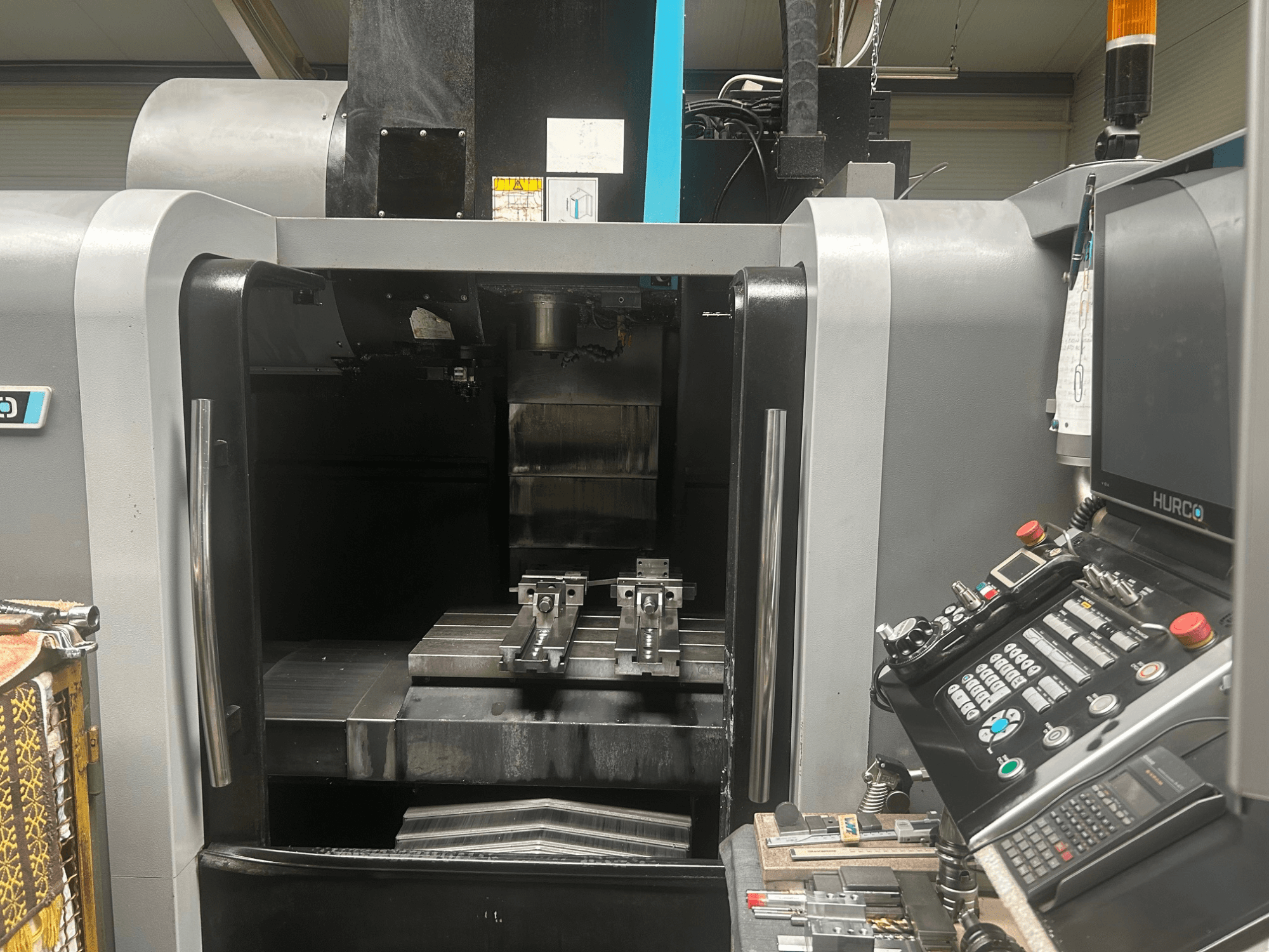 Vista frontale del centro di lavoro CNC Hurco, che mostra il portautensili, il pezzo di metallo e il pannello di controllo con pulsanti e display.