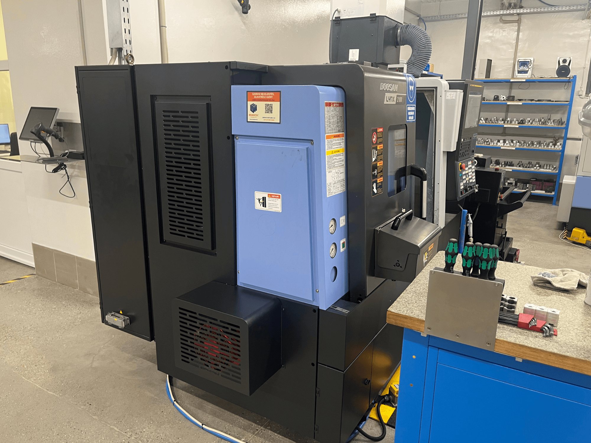 Tornio Doosan Lynx 2100, vista laterale, con pannello di controllo, sistema di ventilazione e utensili su un banco di lavoro.