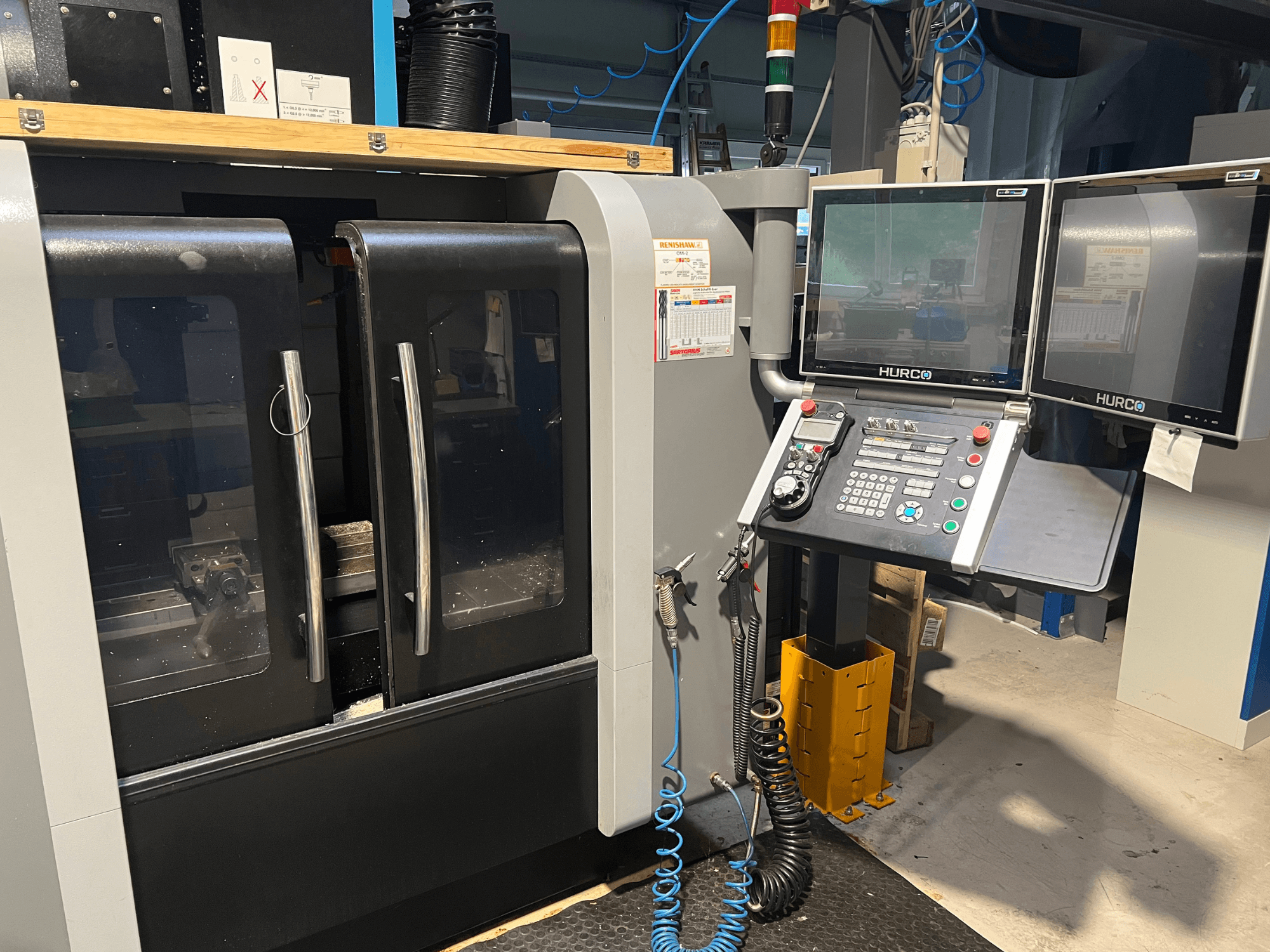 Vista frontale della macchina CNC Hurco con doppio monitor e pannello di controllo. Presenta porte trasparenti e area di lavorazione visibile all'interno.