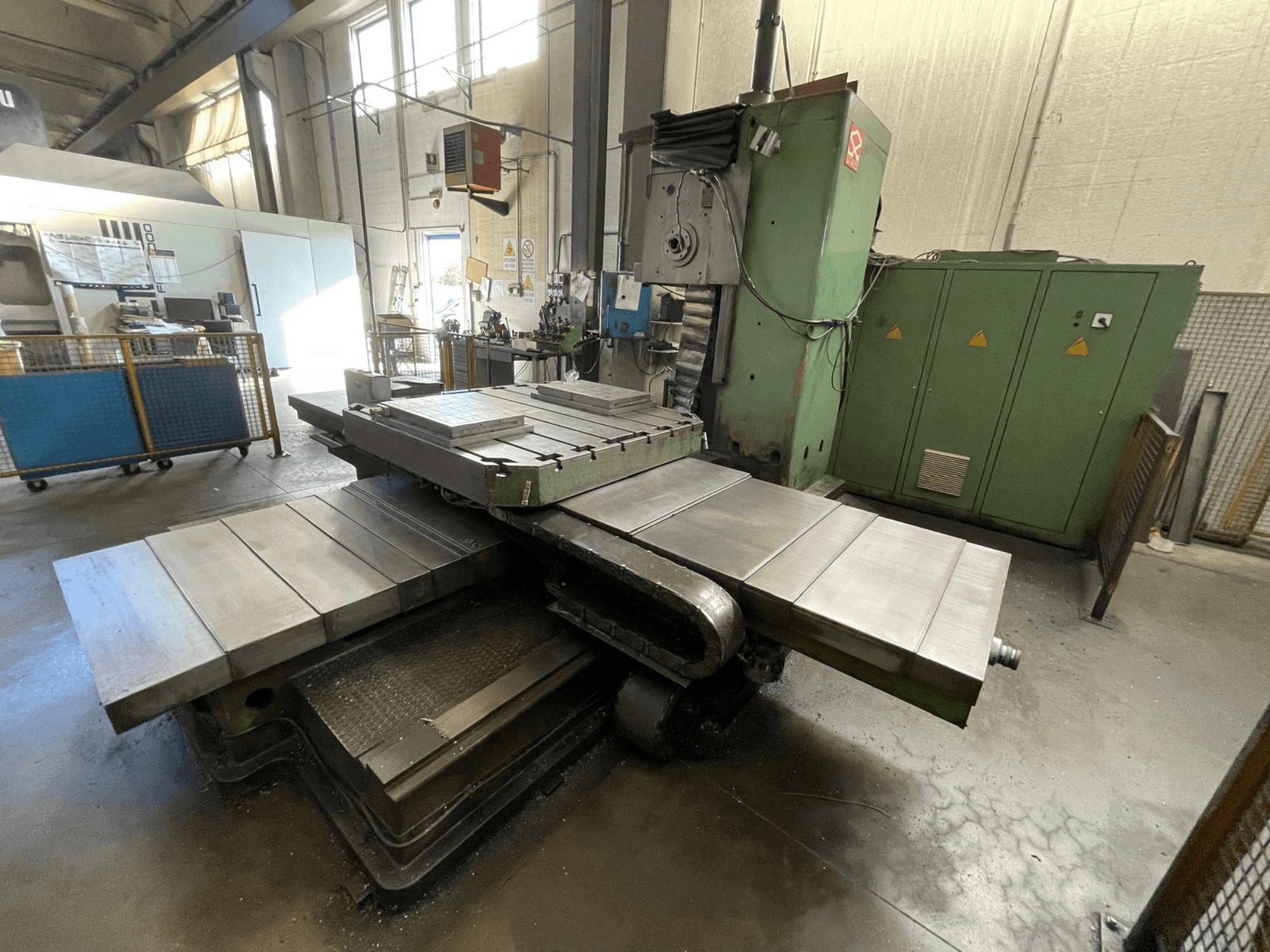 Grande fresatrice industriale con corpo verde, vista laterale, con molteplici superfici piane e armadietti di stoccaggio sullo sfondo.