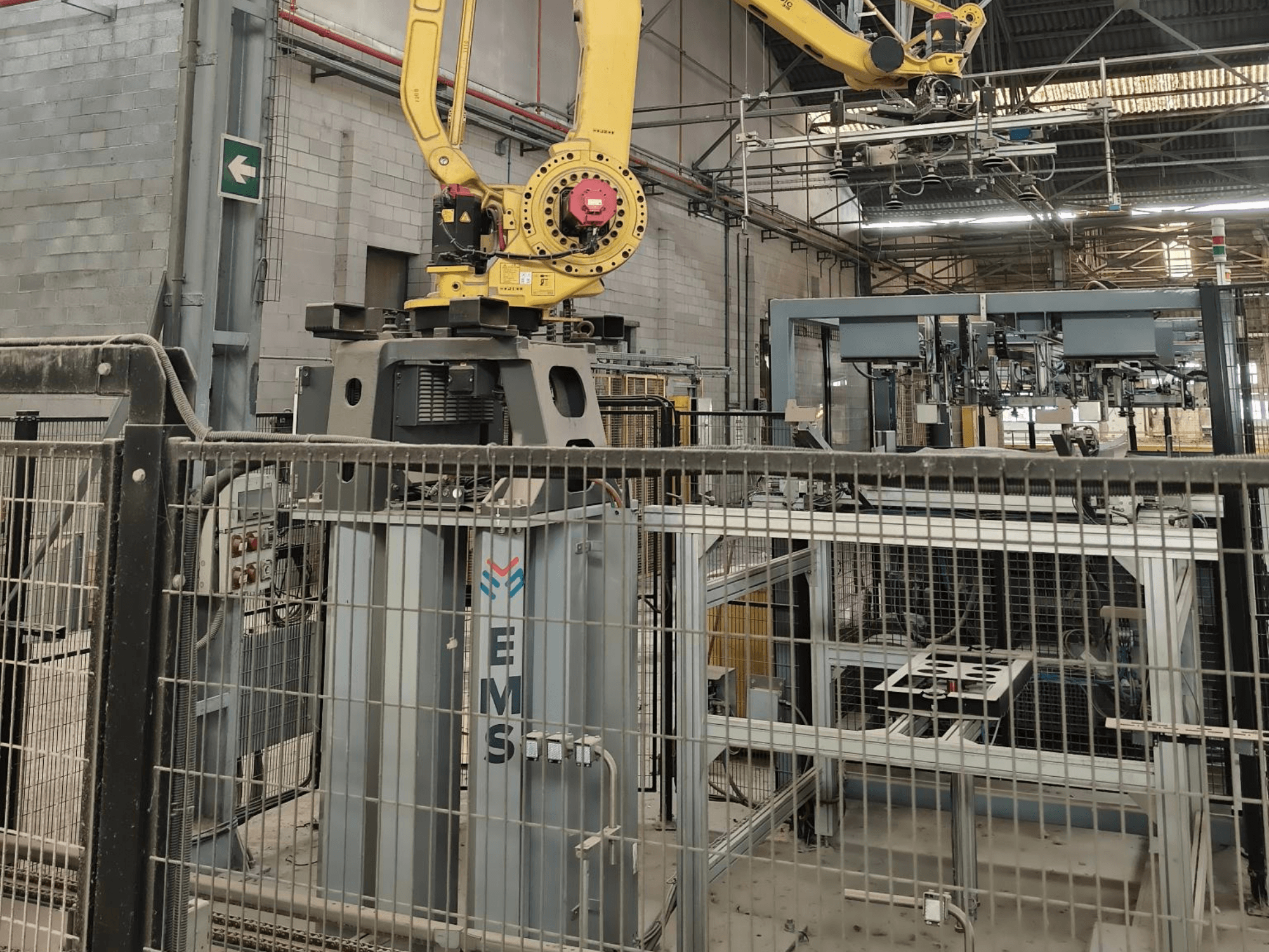 Vista frontale del braccio robotico FANUC M-410iC/315 posizionato sopra un utensile EMS, con attrezzature industriali e recinzioni sullo sfondo.