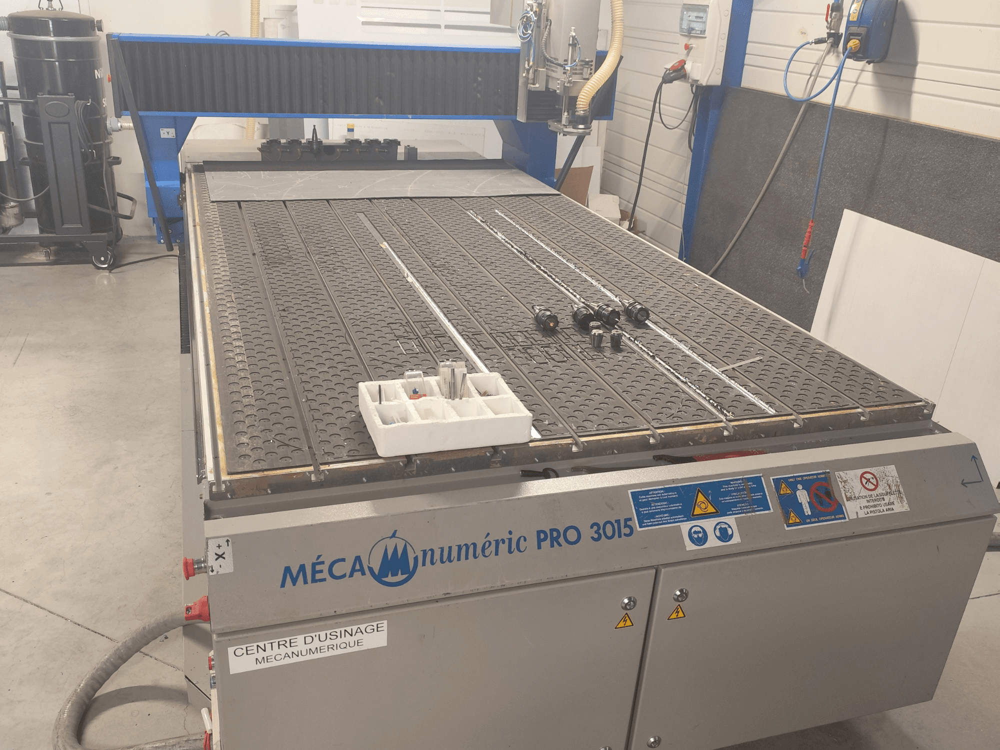 Vista frontale della fresatrice CNC MECANUMERIC Pro 3015, con una dettagliata immagine del piano di lavoro, del pannello di controllo e degli accessori presenti sul tavolo.