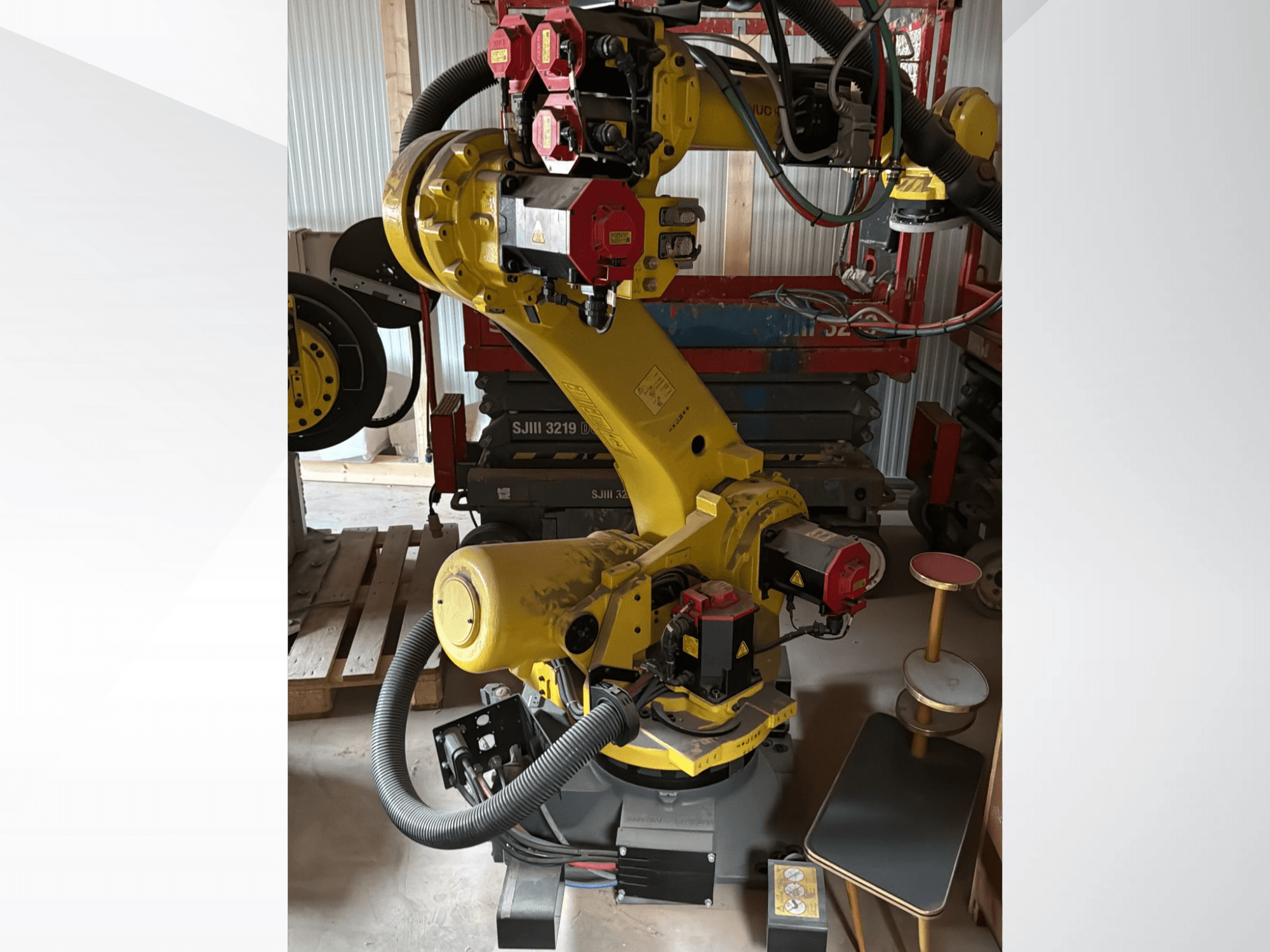 Braccio robotico industriale giallo con controller rossi, vista frontale; dotato di più articolazioni e tubi, ambientato in un cantiere.