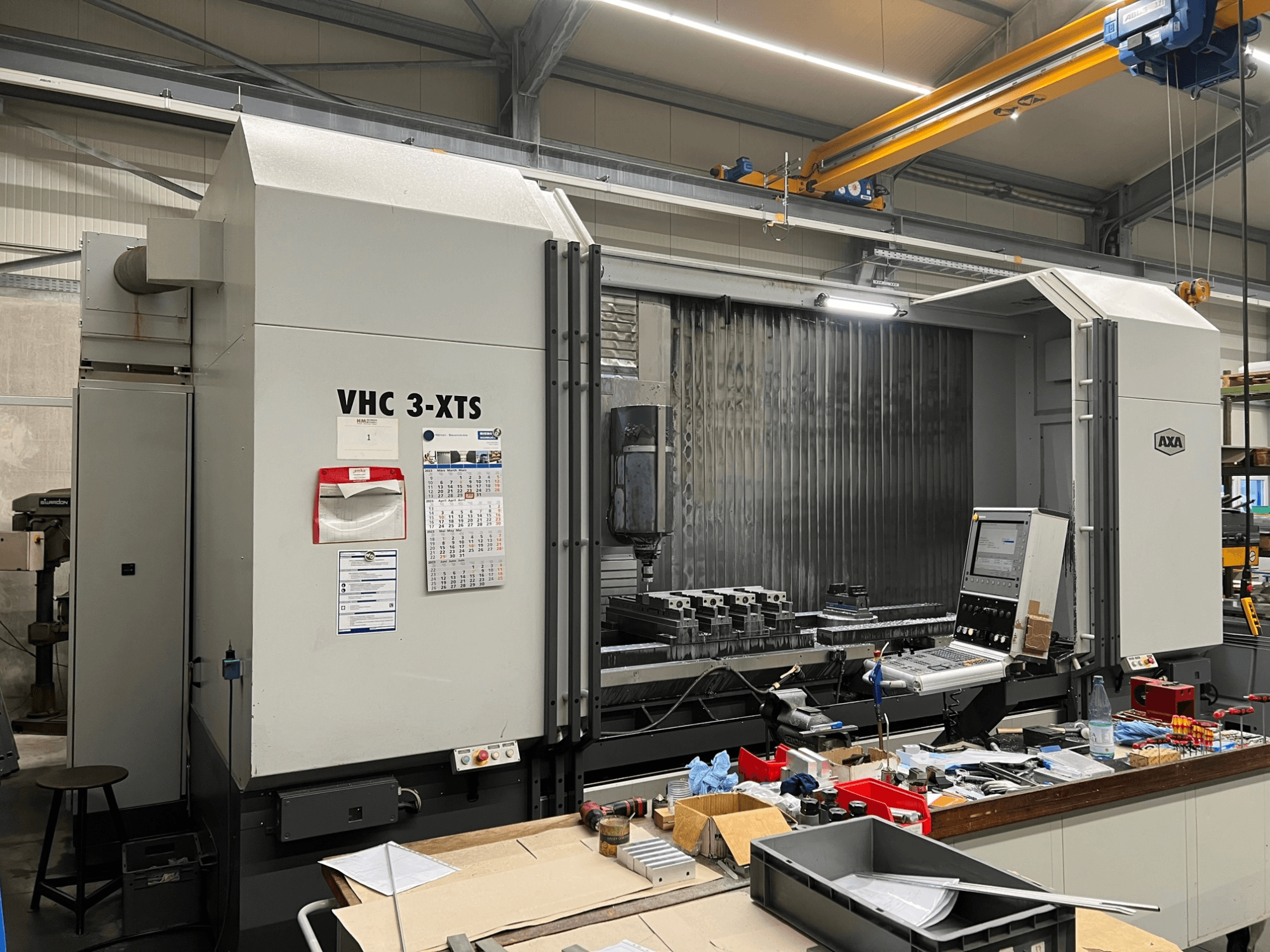 Macchina CNC AXA VHC 3-XTS in vista laterale, con pannello di controllo e area di lavoro ingombra di utensili e componenti.