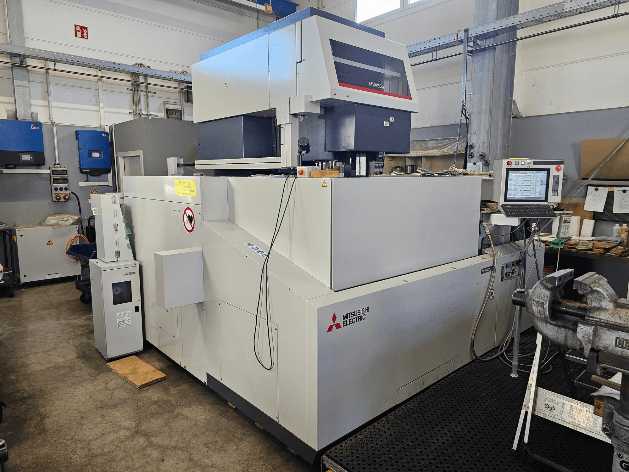 Macchina per elettroerosione a filo Mitsubishi MV4800 in un'officina, vista laterale che mostra il pannello di controllo, le etichette di sicurezza e le attrezzature circostanti.