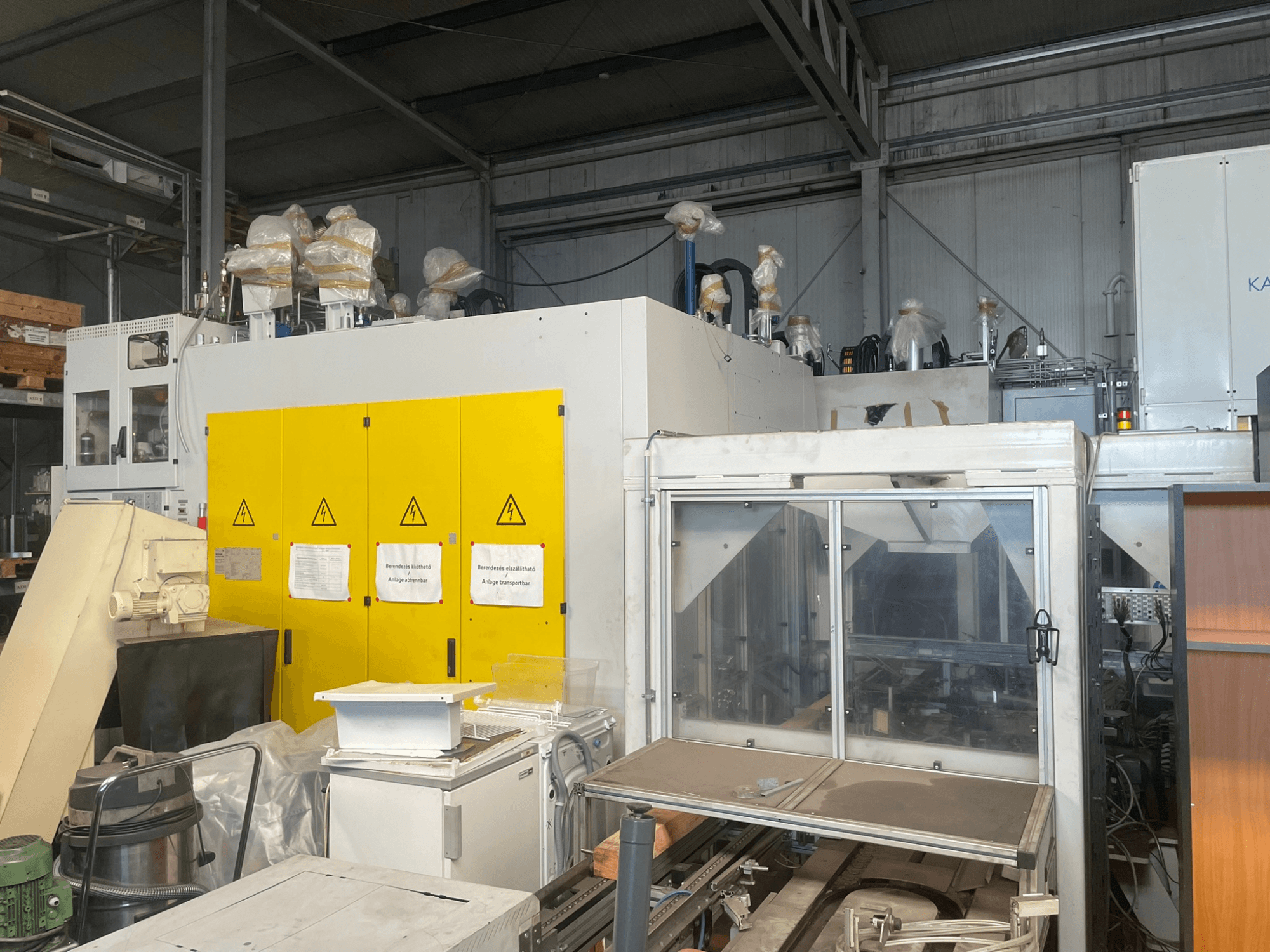 Macchinario industriale con pannello di controllo giallo, vista laterale, con etichette di avvertimento e coperture in plastica, in un magazzino.