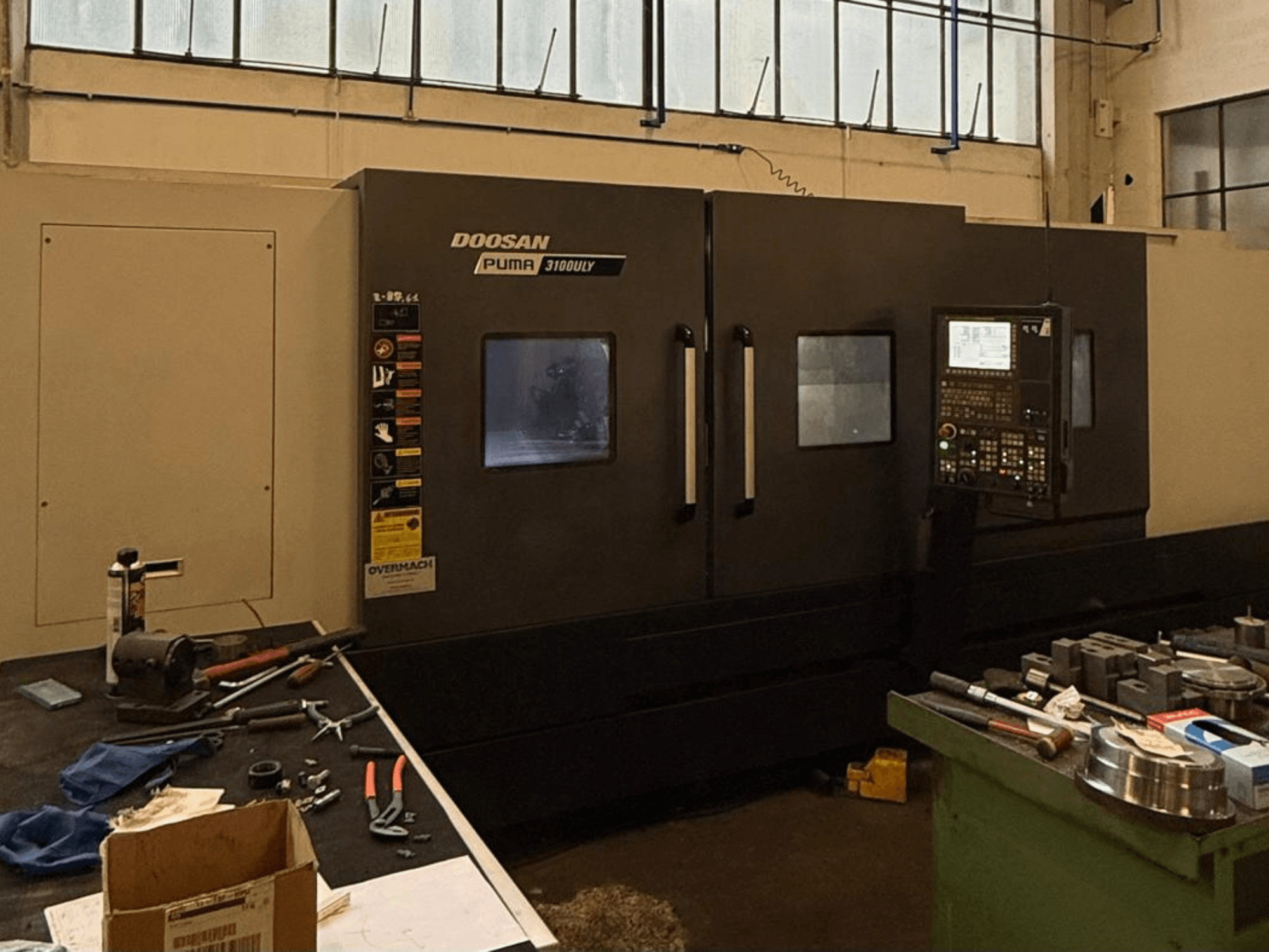 Tornio CNC Doosan Puma 3100LY in un'officina, vista laterale, con pannello di controllo e utensili visibili sul banco di lavoro.