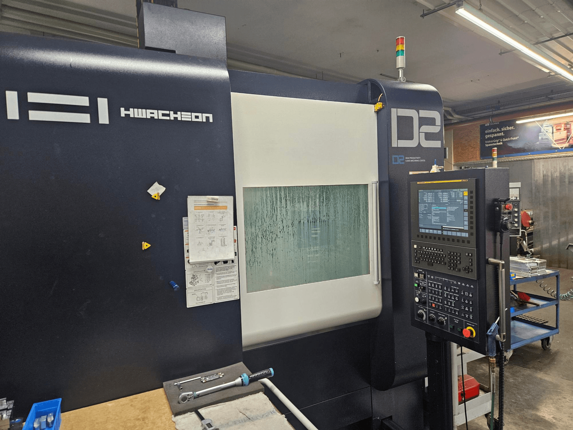 Centro di lavorazione verticale HWACHEON D2-5AX 12k, vista frontale che mostra il pannello di controllo e la finestra di osservazione appannata.