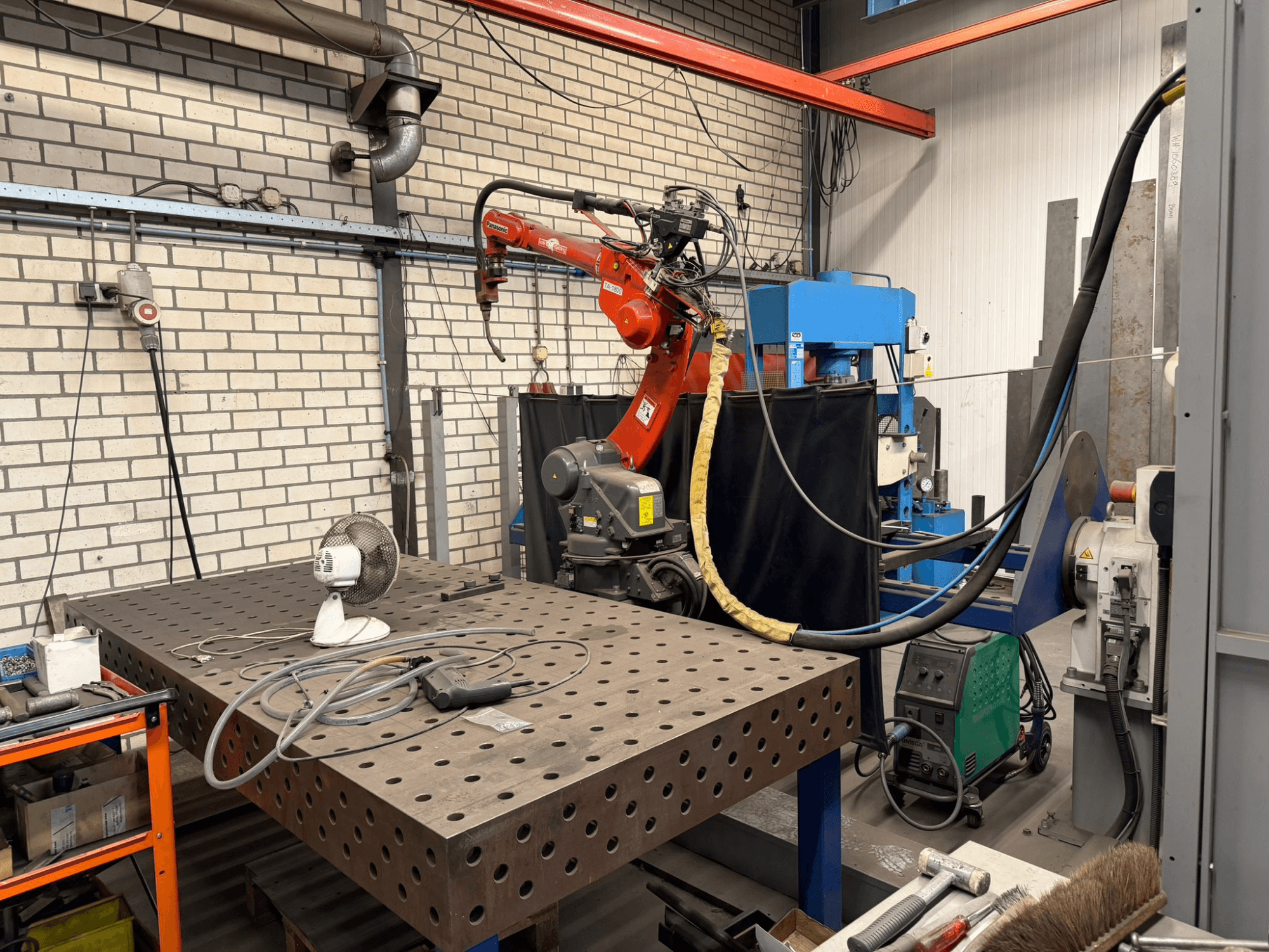 Vista frontale di un robot di saldatura PANASONIC TA-1800 su una postazione di lavoro, con ventilatore e attrezzatura di saldatura visibili.