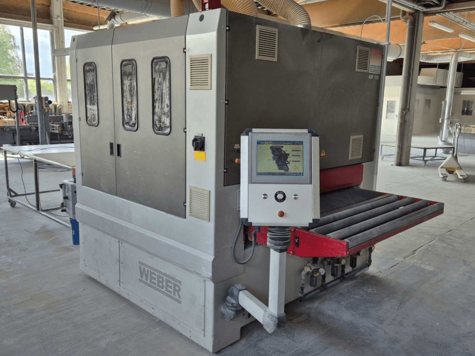 Levigatrice a nastro WEBER KSF-3-1350, vista laterale che mostra i comandi, il pannello di alimentazione e il nastro di levigatura. Ambiente industriale con macchine visibili.