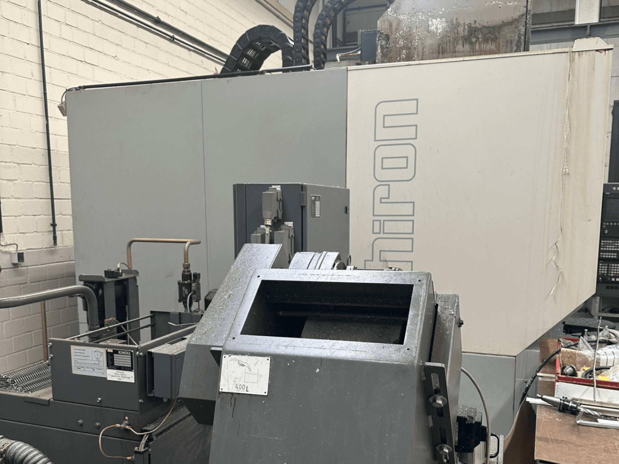 MARCA: Chiron; Vista frontale di una macchina industriale a controllo numerico con esterno grigio, pannello di controllo visibile e componenti del macchinario a fuoco.