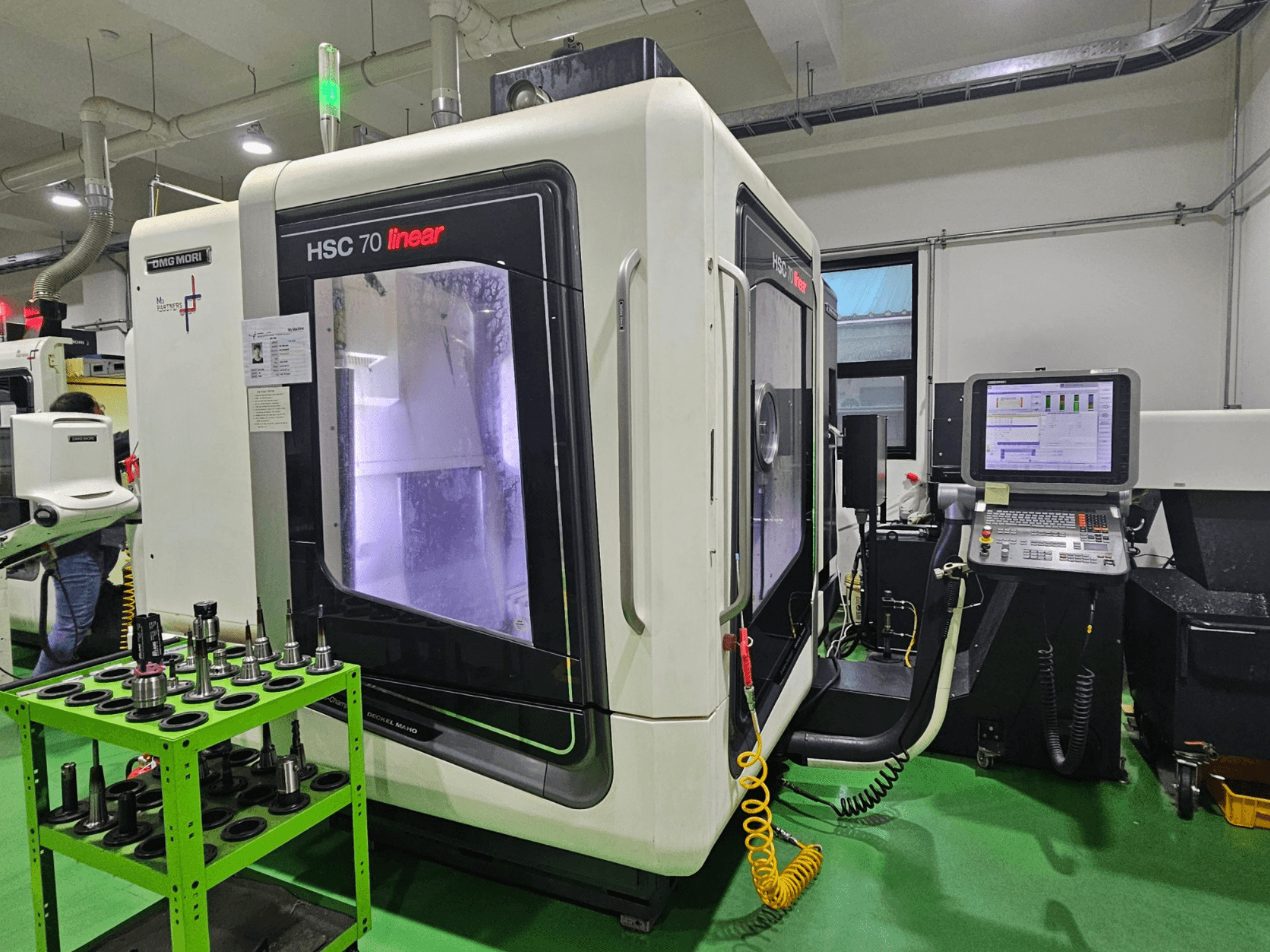 Centro di lavoro universale lineare DMG MORI HSC 70, vista frontale, con portautensili su un tavolo verde e pannello di controllo visibile.