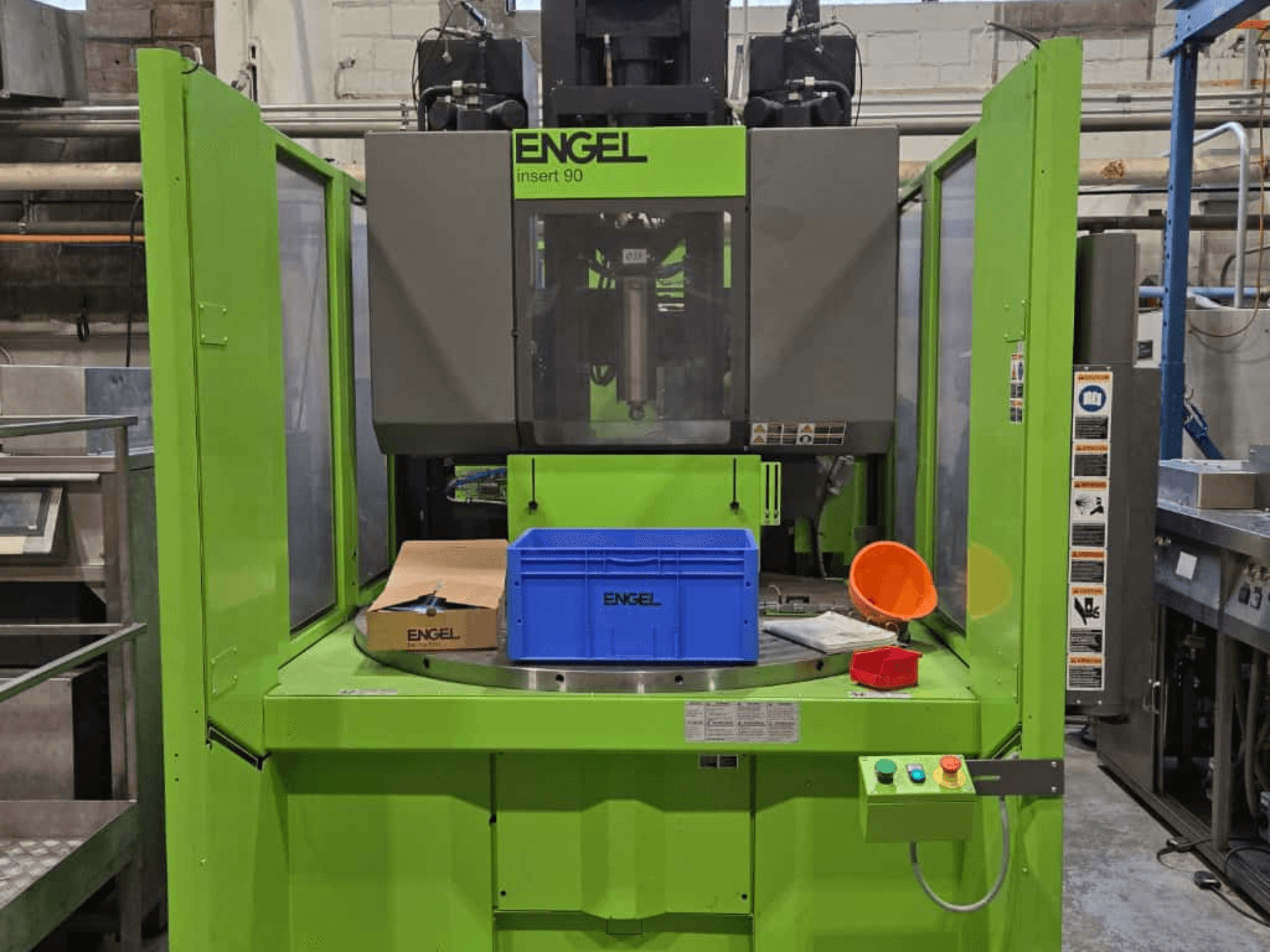 Vista frontale della macchina di stampaggio a iniezione ENGEL Insert 90 verde, con contenitore blu e pannello di controllo con pulsanti.