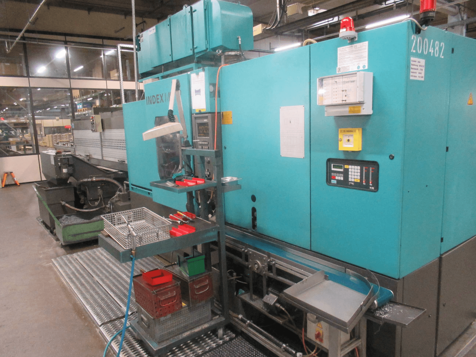 Macchina INDEX turchese in fabbrica, vista laterale; dispone di pannello di controllo, magazzino utensili e trasportatore. Sono visibili bidoni, tubi e allarmi.