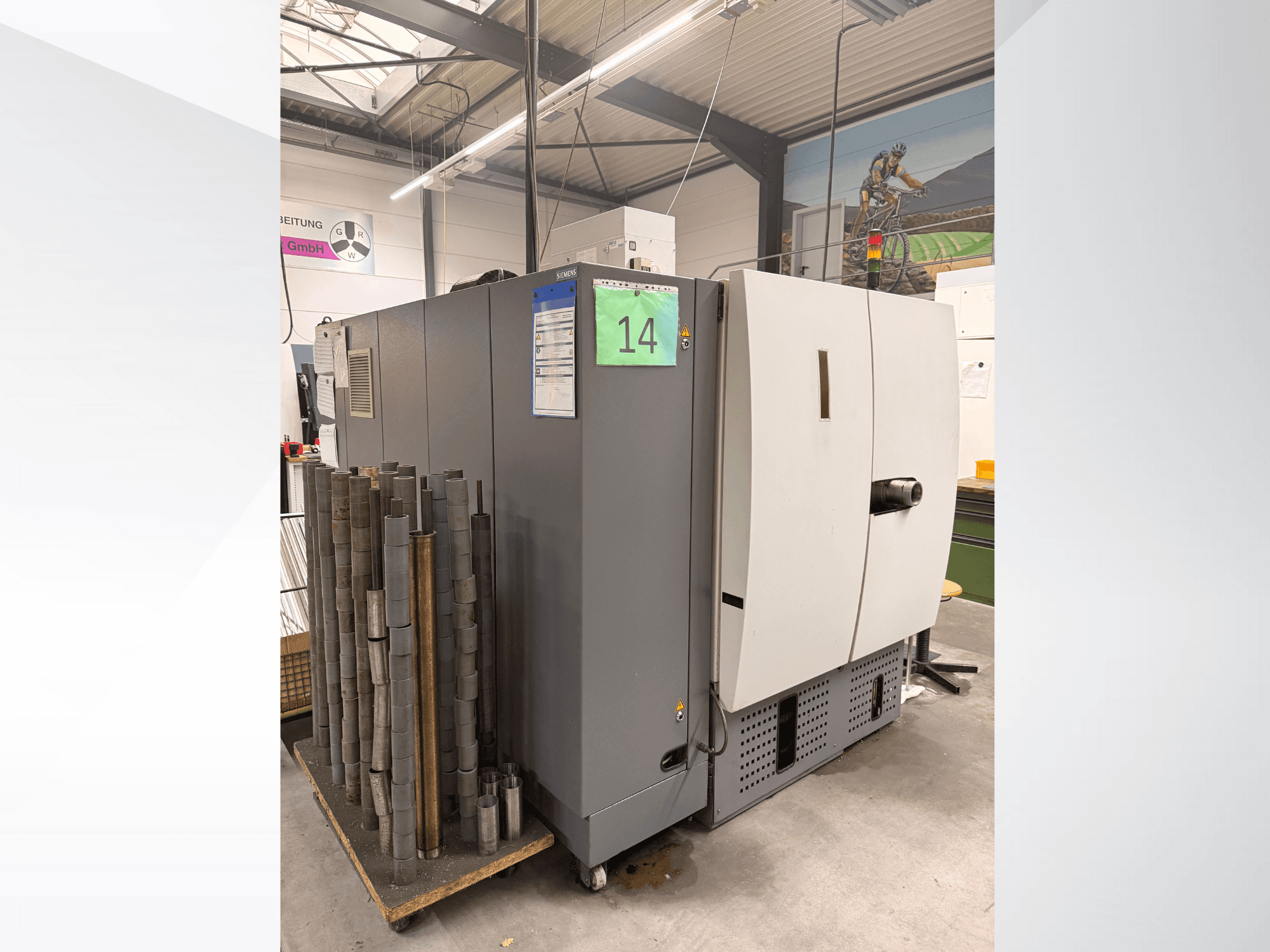 Macchina industriale grigia di Siemens, vista laterale, con accanto uno stock di pezzi metallici cilindrici e le etichette delle attrezzature visibili.