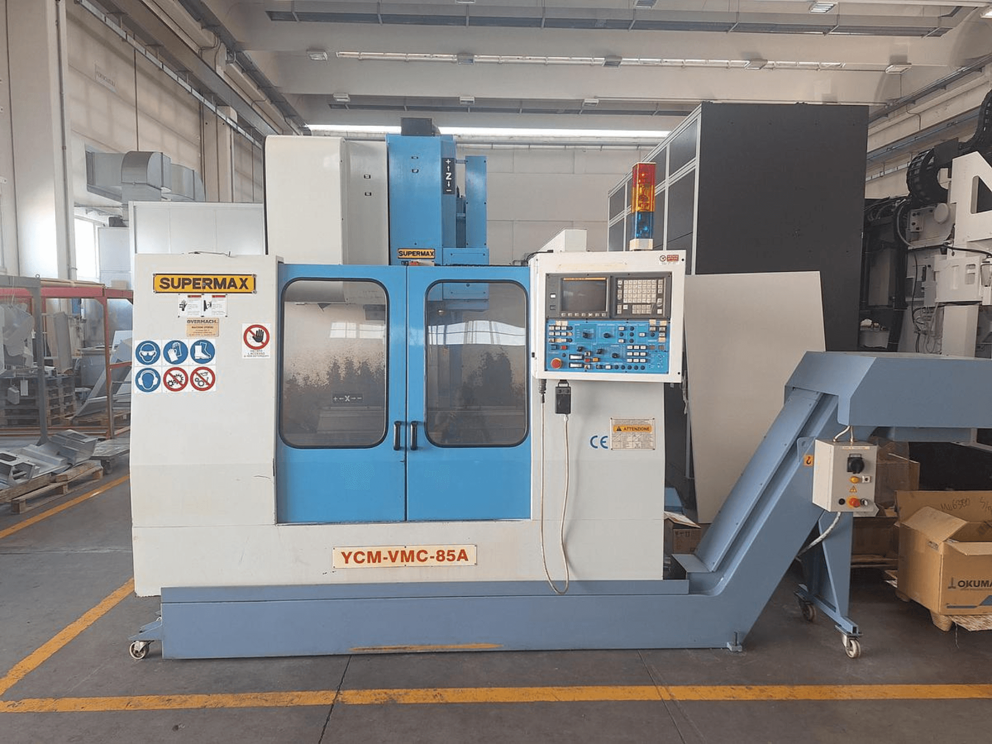 Centro di lavorazione verticale SUPERMAX YCM-VMC-85A in uno stabilimento, con pannello di controllo e adesivi di sicurezza, vista frontale.