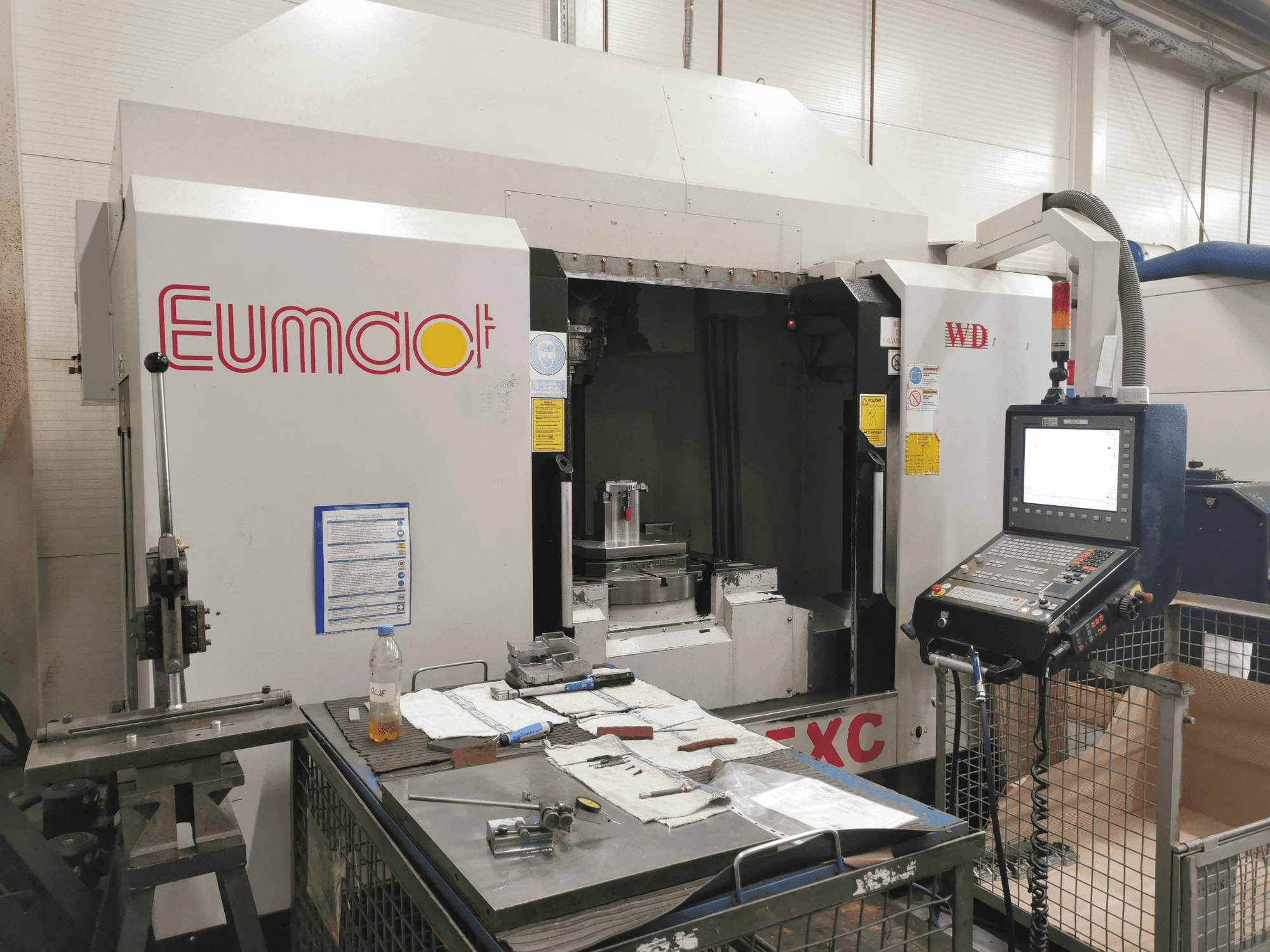 Vista frontale di una macchina CNC Eumdaot con pannello di controllo, utensili e una superficie di lavoro ingombra di componenti e materiali.