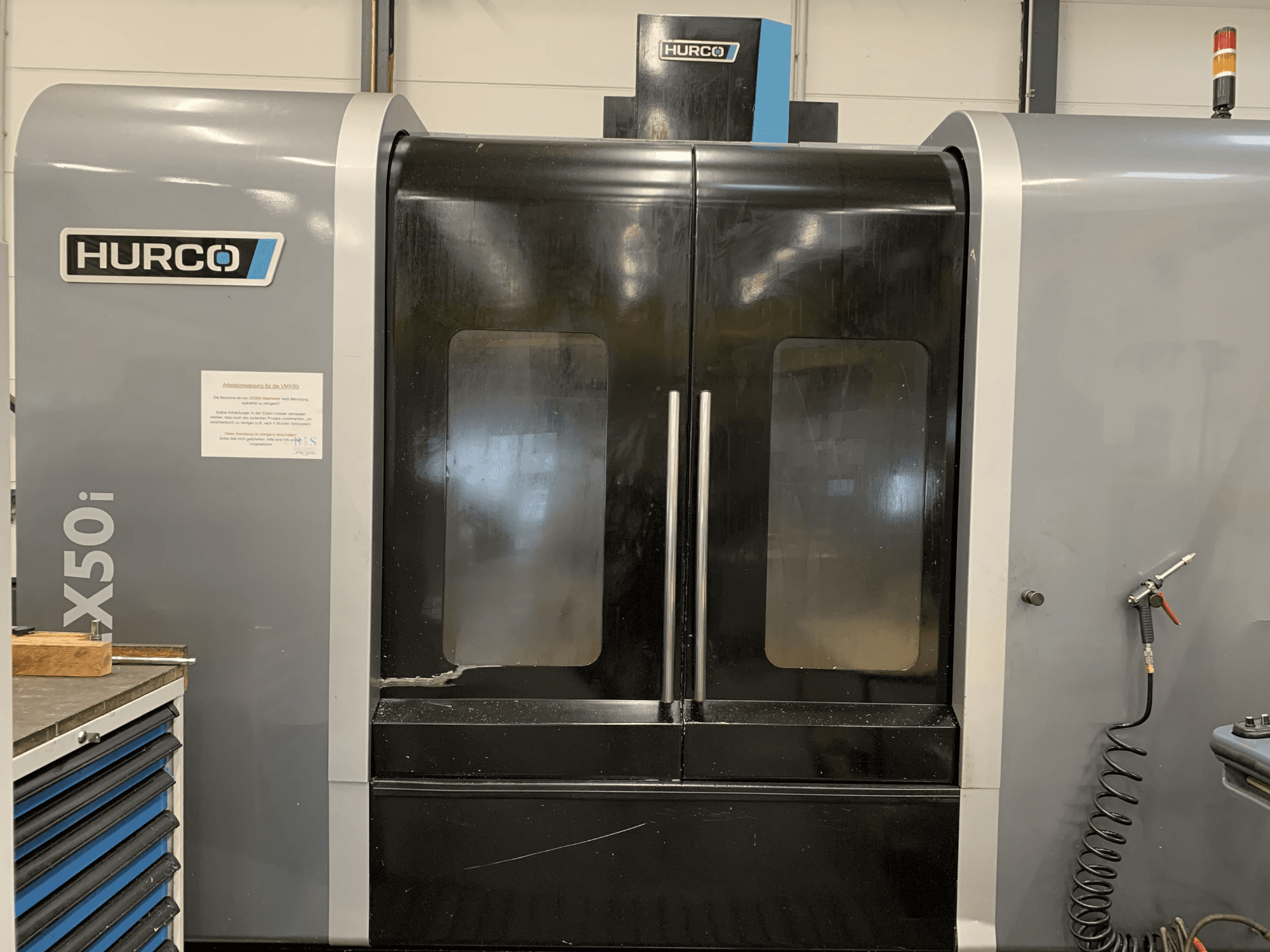 Vista frontale di una macchina CNC Hurco VX50i a doppia porta, con comandi tecnici e cartello di manutenzione sul lato.