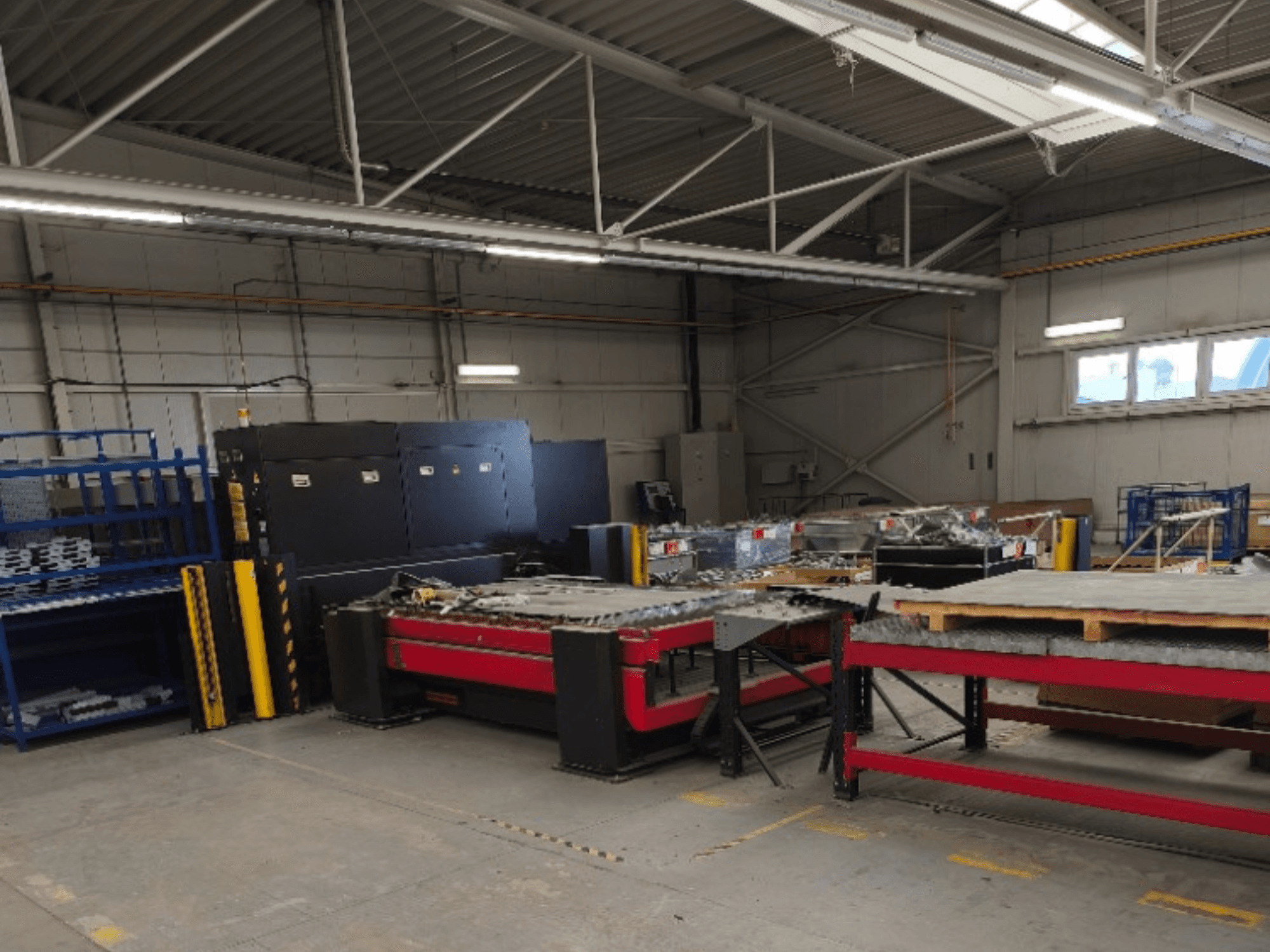 Macchina per il taglio laser a fibra AMADA LCG 3015 AJ in un'officina, vista frontale con scaffali di stoccaggio e materiali sullo sfondo.