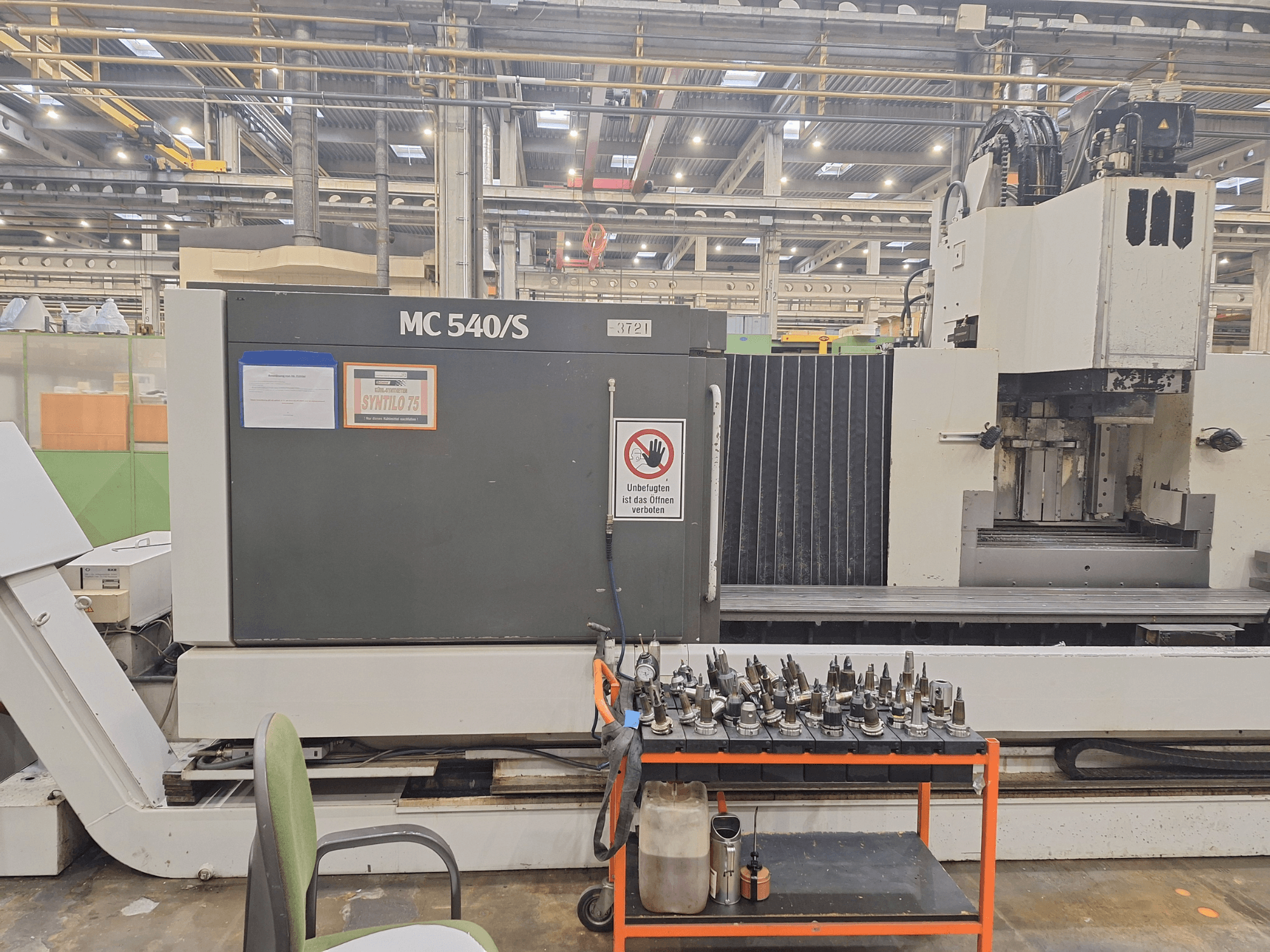 Centro di lavoro verticale STAMA MC 540S in un'officina, vista laterale con i portautensili su un carrello vicino.