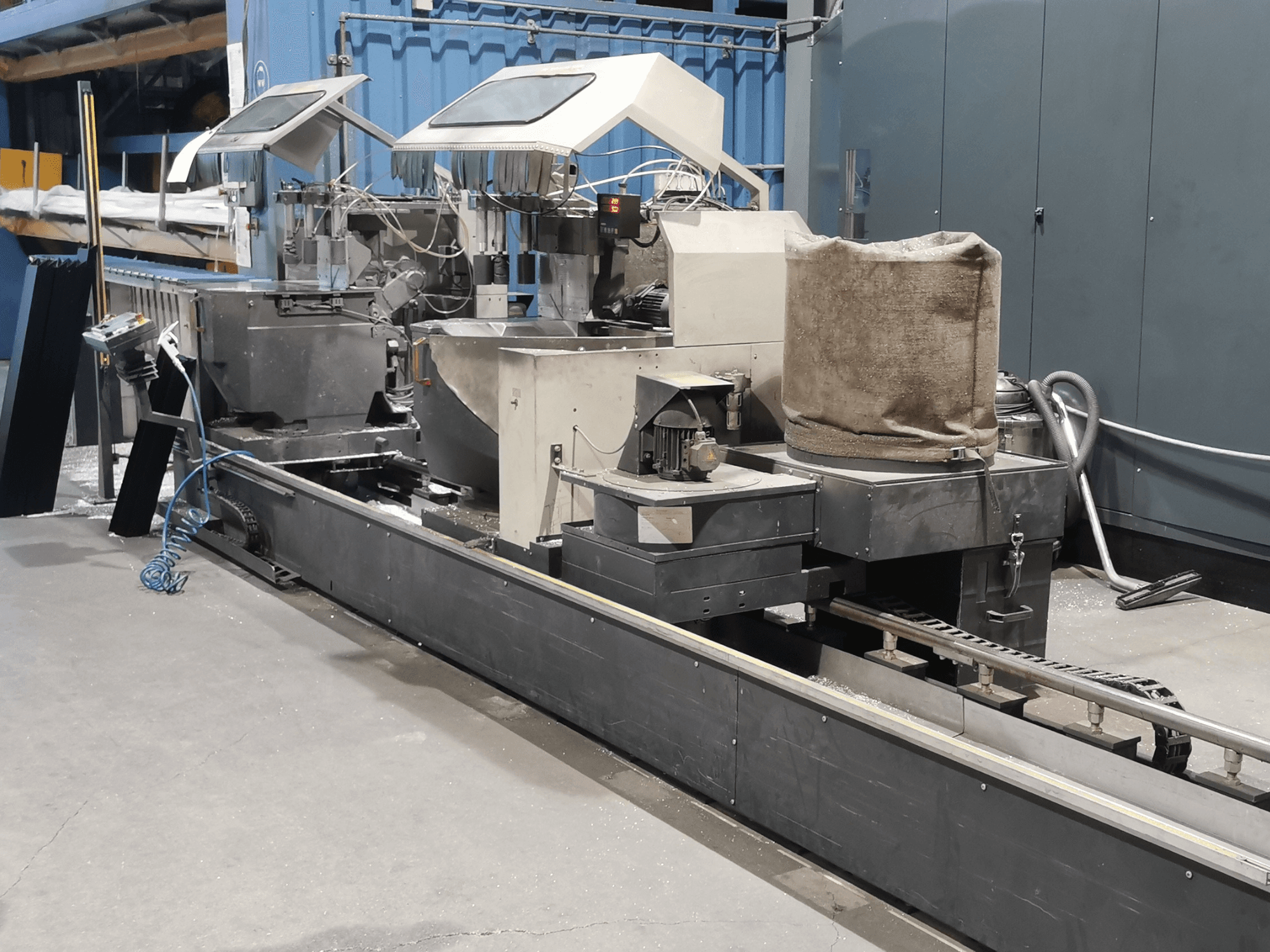 Macchina di lavorazione industriale vista di lato, con tramogge, nastro trasportatore e pannello di controllo, in un ambiente di fabbrica.