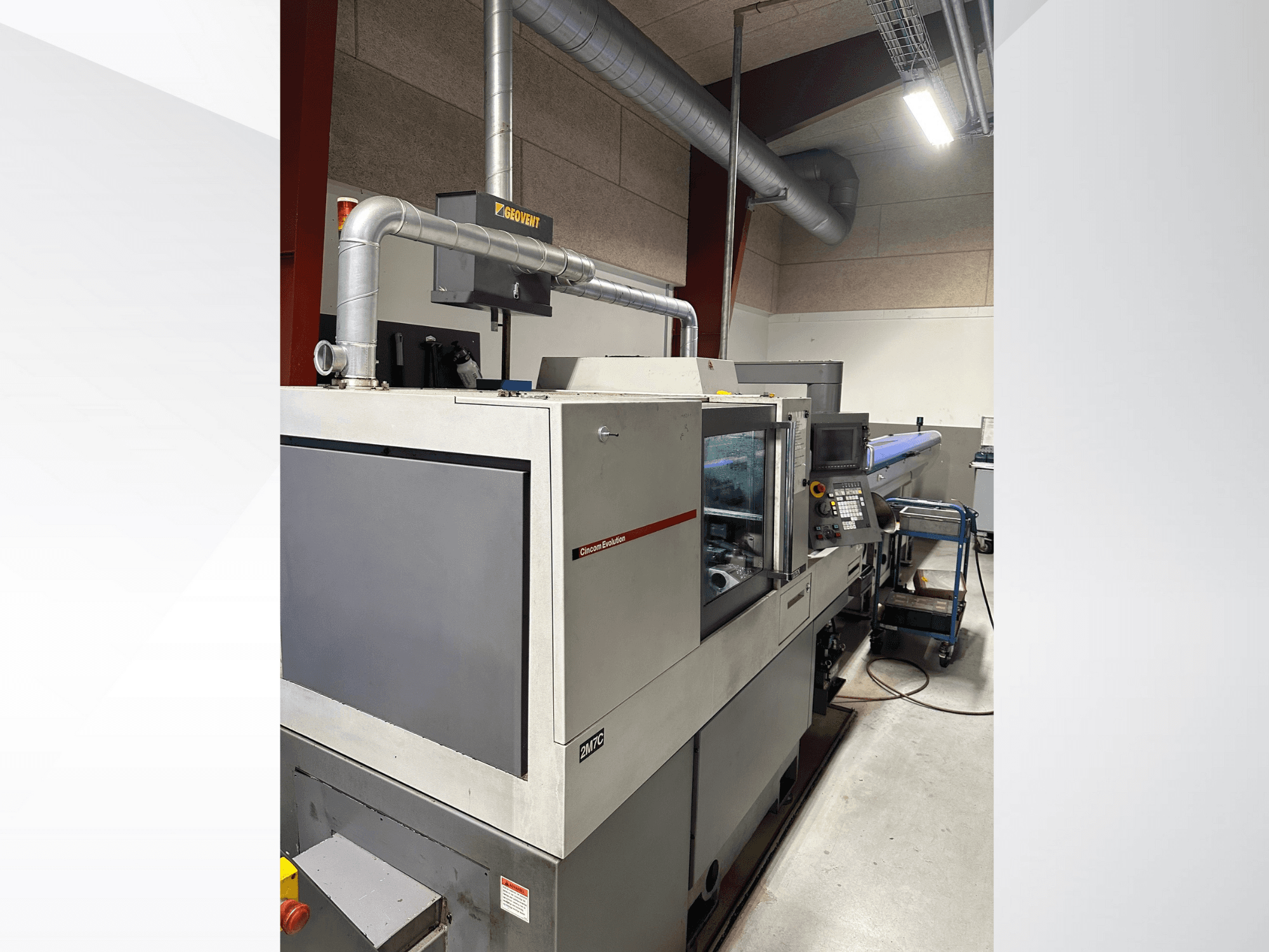 Macchinario industriale del marchio MVC, vista laterale con pannello di controllo e canalizzazione visibili, ambientato in una fabbrica.