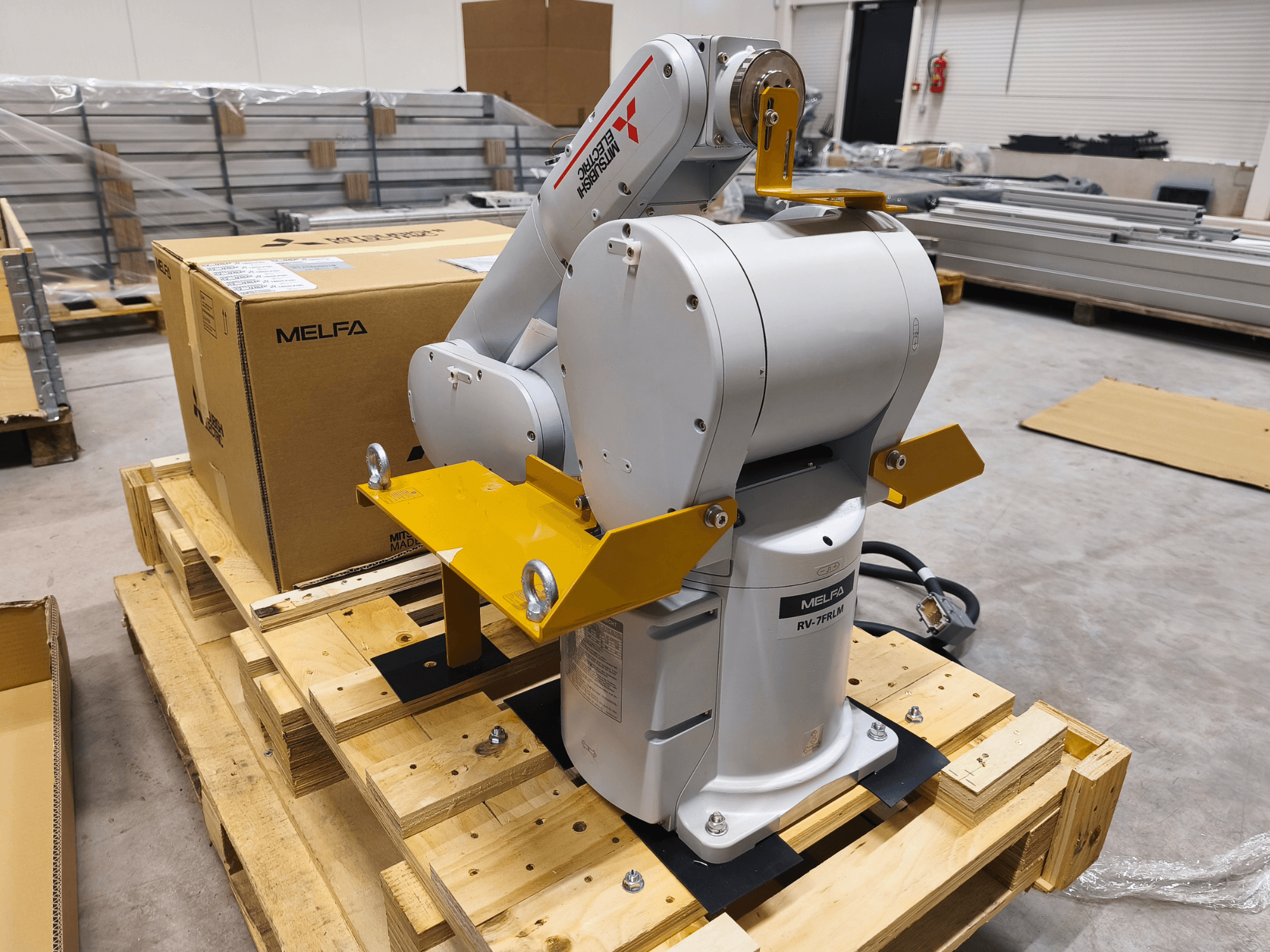 Braccio robotico industriale MELFA RV-7FLM su pallet di legno, vista frontale, con staffe di supporto gialle e scatola di imballaggio grigia.