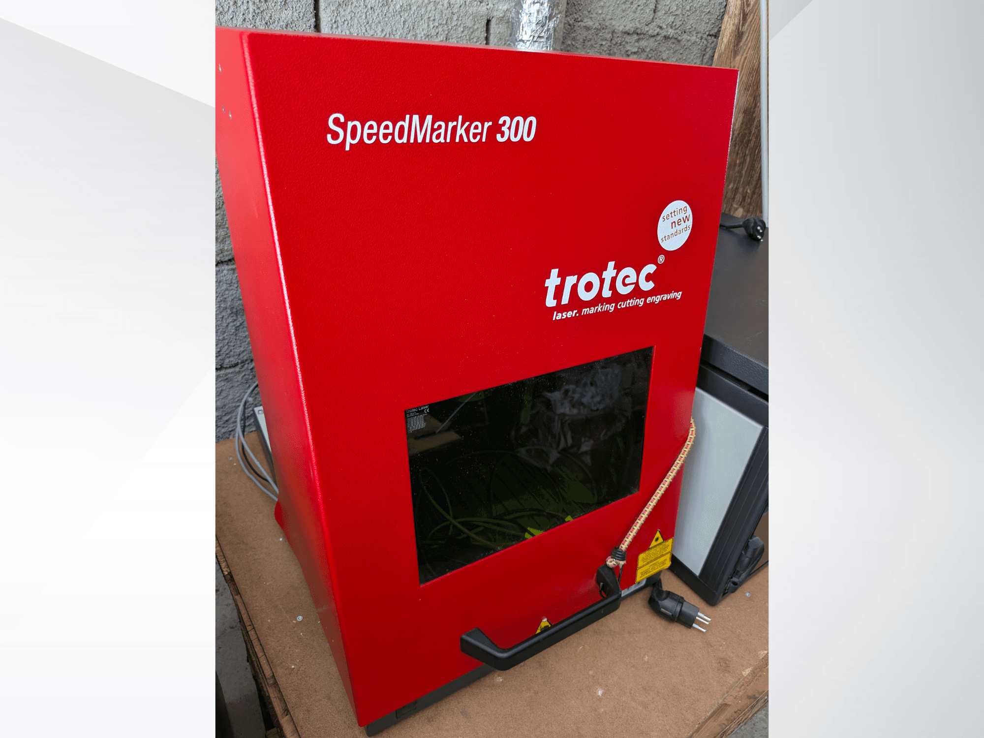 Macchina di marcatura laser Trotec SpeedMarker 300 rossa in vista frontale, con ampia finestra di visualizzazione e connessione di alimentazione.