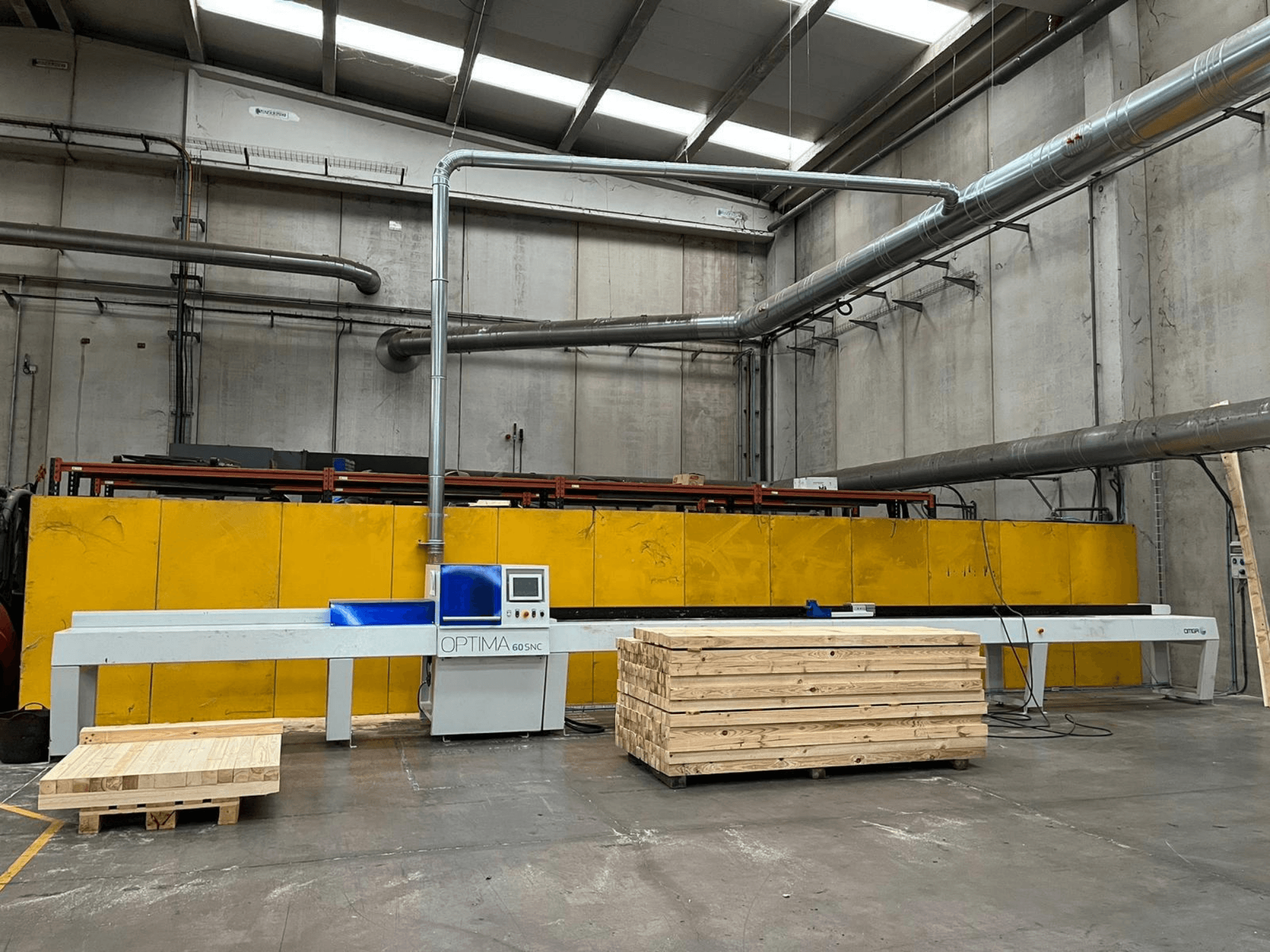Vista frontale della sega trasversale ottimizzata OPTIMA 60SNC 6200 CE, con pallet di legno e barriere di sicurezza gialle sullo sfondo.