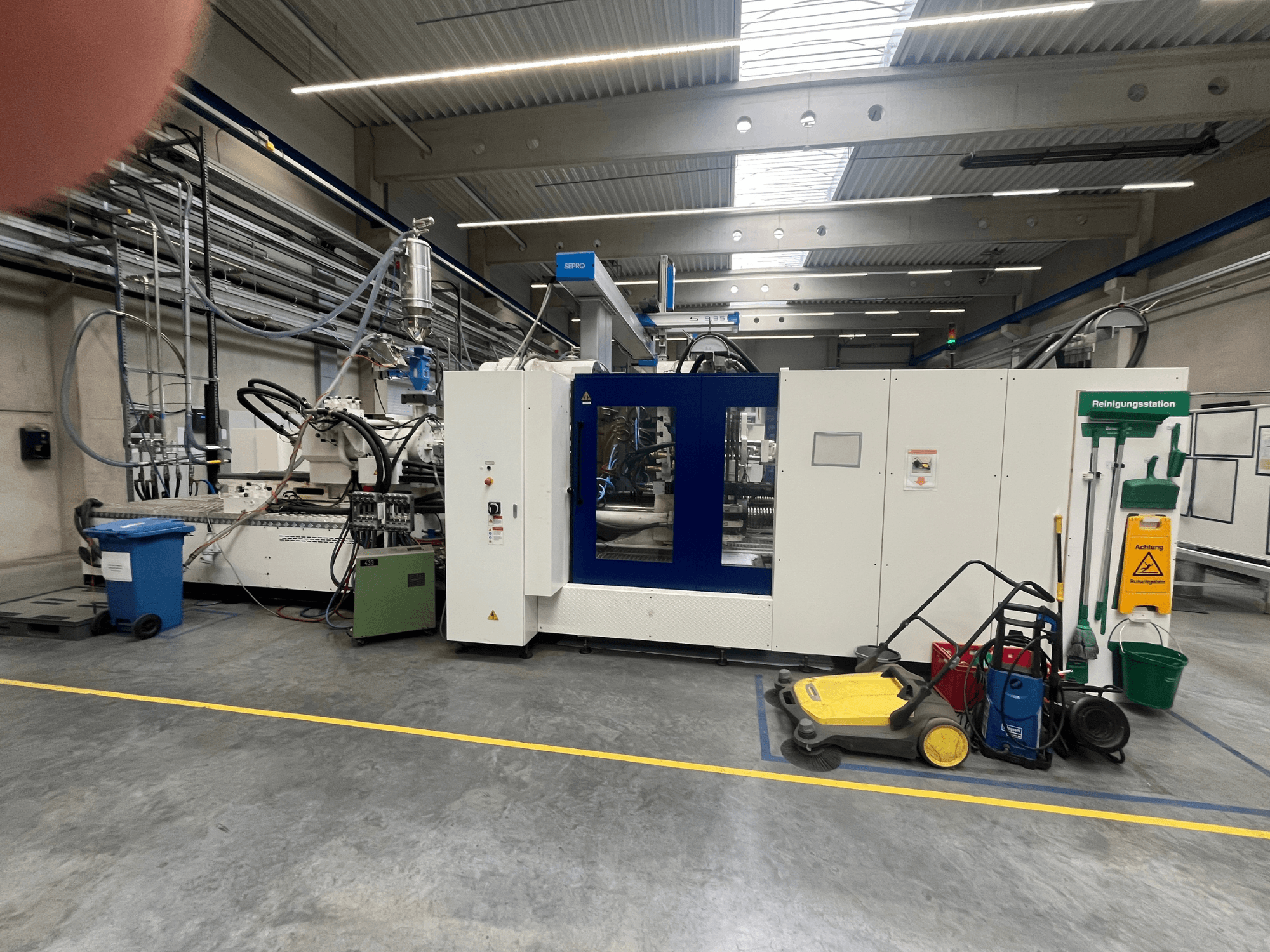 Macchina industriale in una fabbrica, vista frontale, con pannelli blu e bianchi, tubi e attrezzature di pulizia nelle vicinanze.