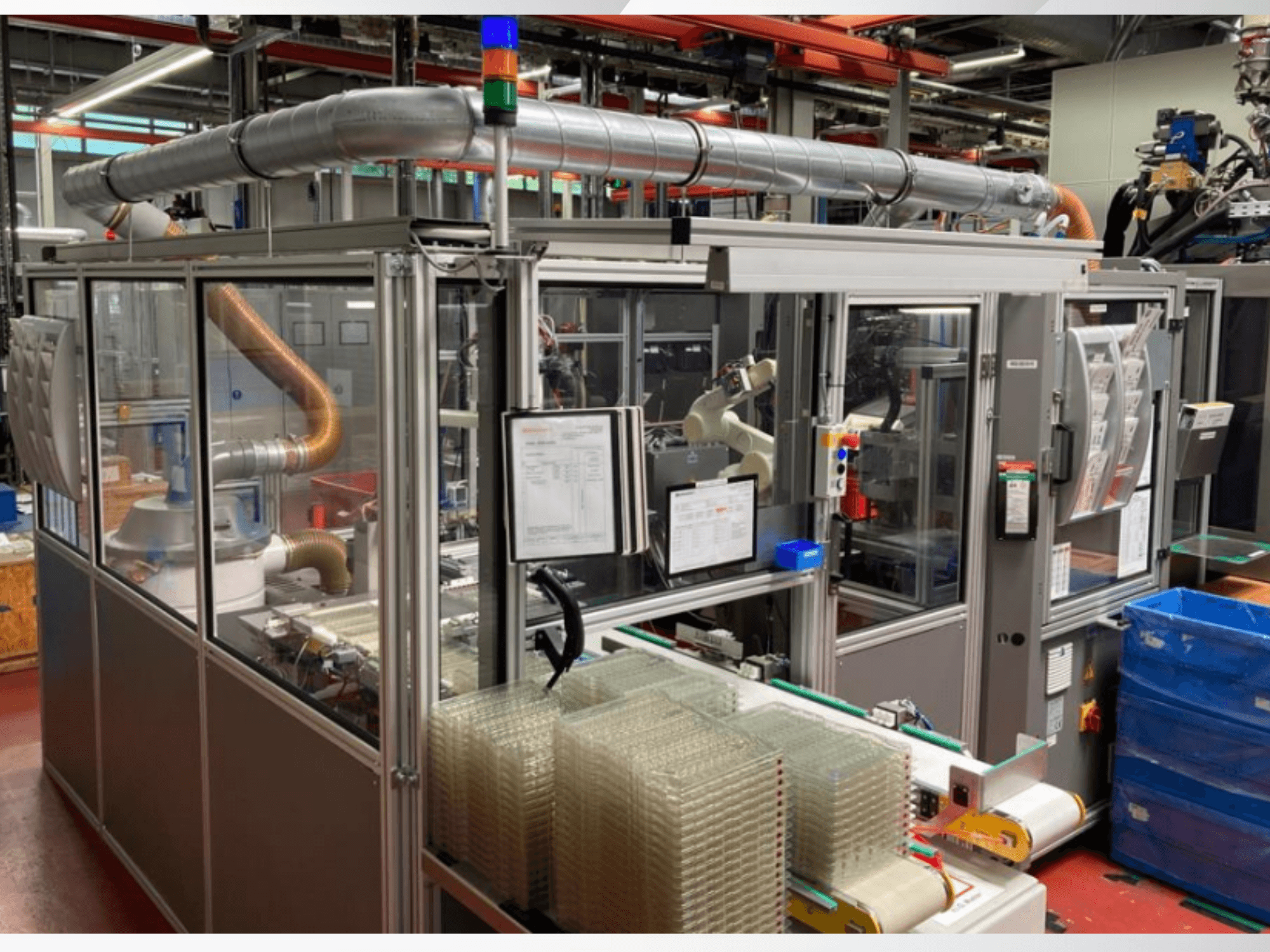 Vista frontale di una macchina automatizzata con tubi metallici, pannello di controllo e contenitori di plastica trasparente impilati su un pavimento di fabbrica.