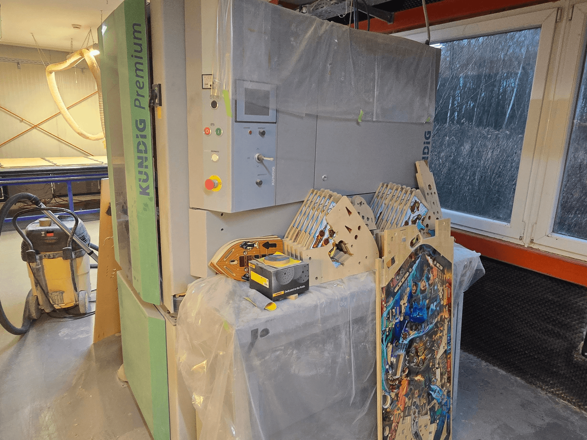 Levigatrice a nastro Kundig Premium-1 1100-C in officina, con pannello di controllo e pezzi di legno, vista laterale, con aspiratore di polvere sullo sfondo.